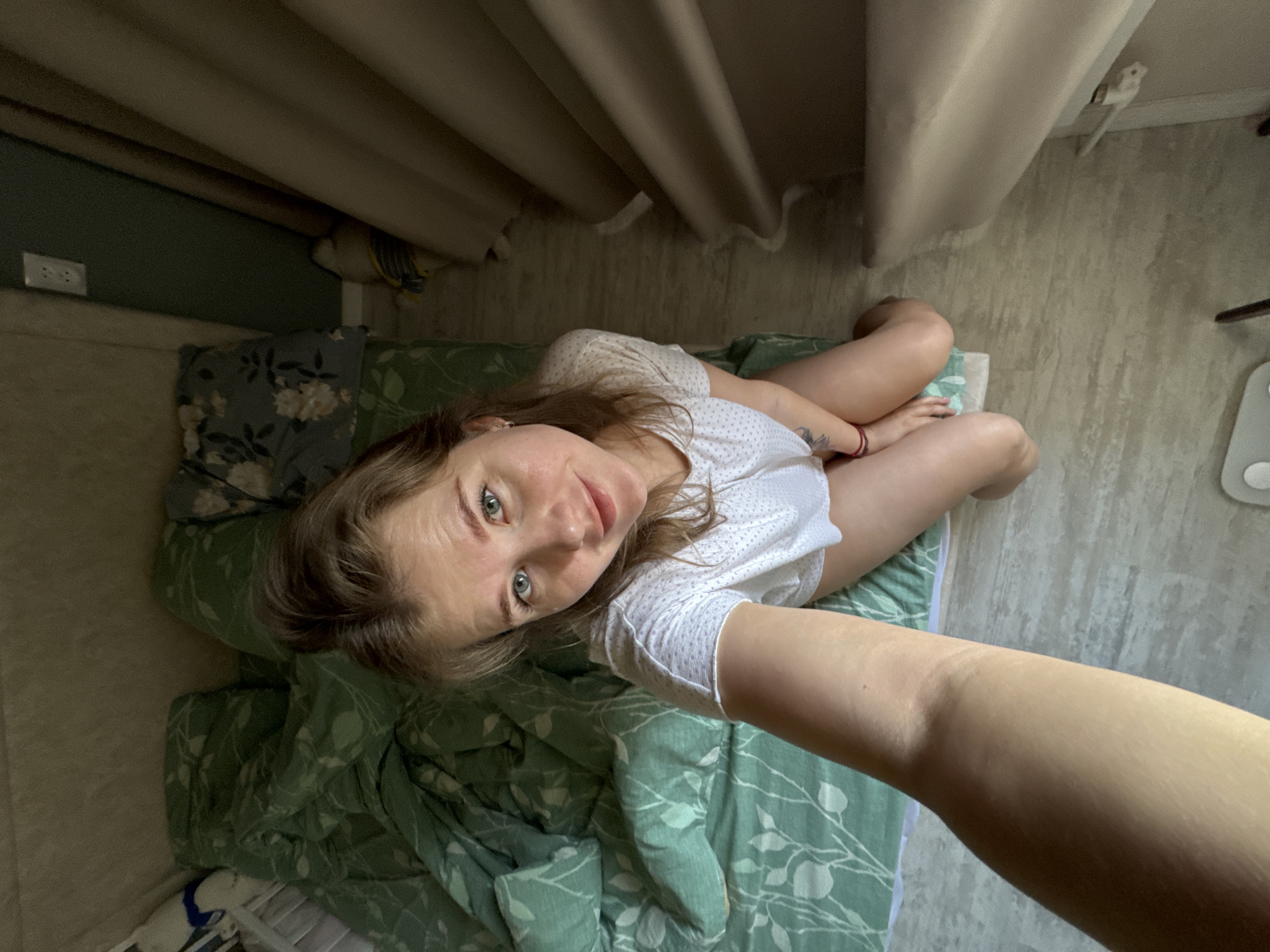 A medium, high-angle shot shows a woman indoors. The woman has light skin, light brown hair, and blue eyes. She is wearing a white short-sleeve shirt. She is sitting cross-legged on a green bed with a leaf pattern. Her arm, likely holding the camera, extends toward the bottom right corner of the frame.
The background includes neutral-colored curtains, a light-colored wall, and a dark green wall. The location is implied to be in Ферма, Russia.