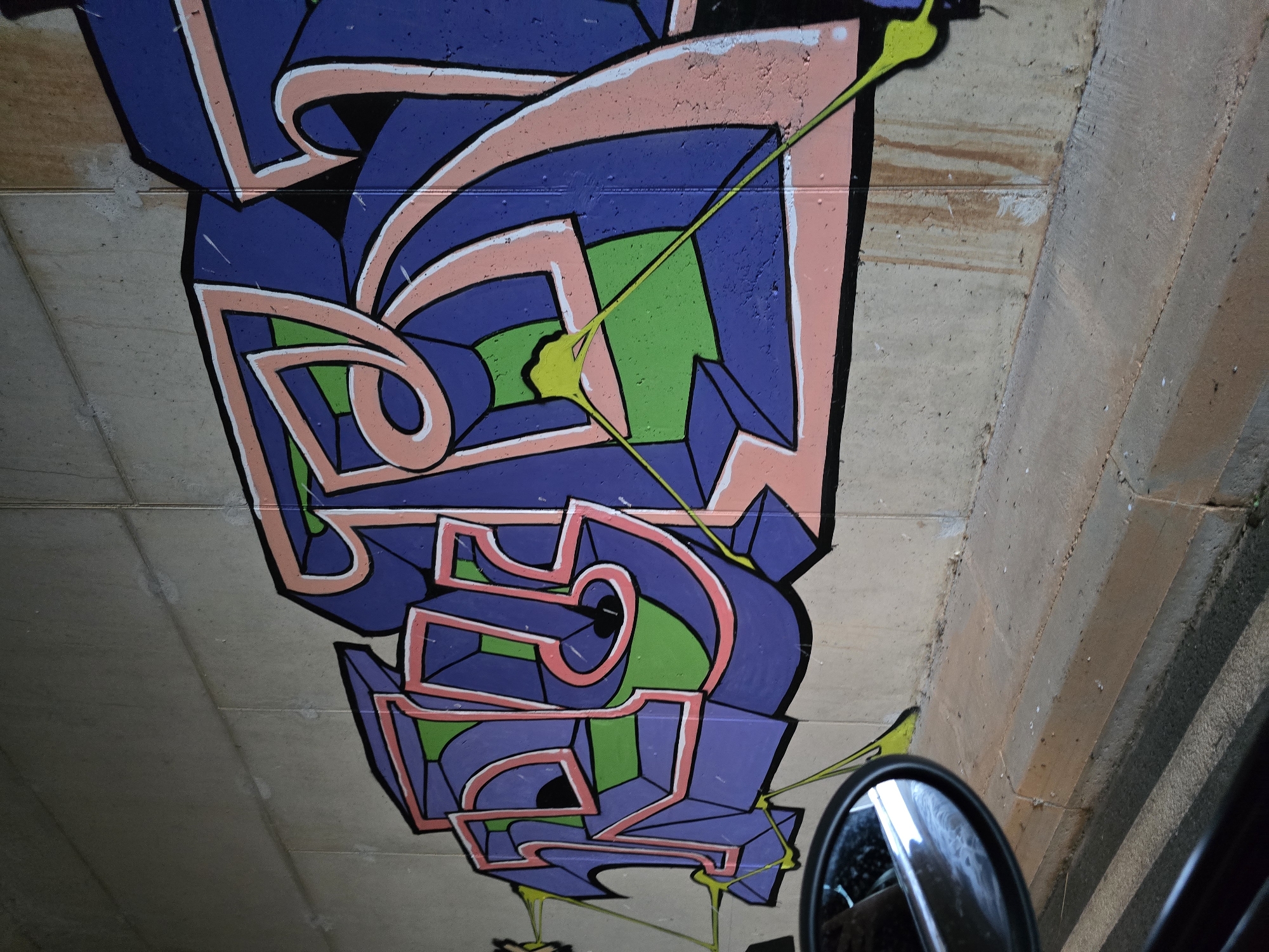 A vibrant graffiti piece covers a concrete wall, likely in an urban underpass or tunnel given the surrounding concrete and the presence of a car's side mirror in the foreground. The graffiti features bold, stylized letters in shades of purple and pink, outlined in white and black. Interspersed within the lettering are sections of bright green. Thin, lime-green lines extend from some of the letters, creating a drip-like effect. The concrete wall itself shows signs of age and weathering, with some water stains and smudges visible. The ground in front of the wall appears to be a combination of concrete and asphalt, suggesting a road or walkway. The car mirror reflects the sky and some of the graffiti. The overall impression is of urban street art in a somewhat gritty, utilitarian setting. The location is specified as Klopot, Montenegro.
