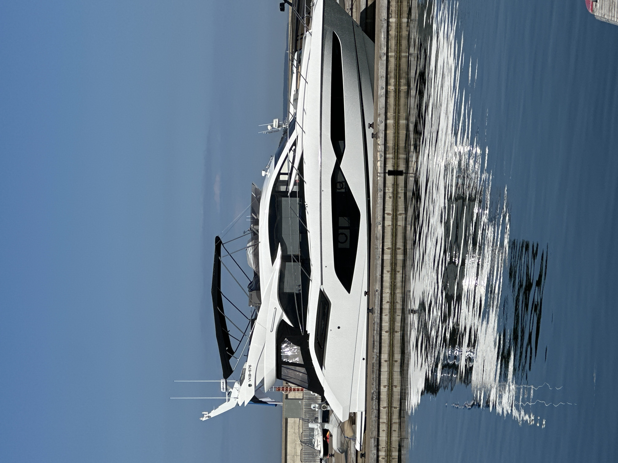 A large, modern white motor yacht is depicted docked in calm, deep blue water under a clear blue sky. The vessel features a sleek, contemporary design with prominent black window lines and hull accents, and a black canvas bimini covering part of its upper deck. The registration number "VKB-576" is visible on the stern, along with a white radar dome (branded "SIMRAD") and two tall white antennas mounted on the hardtop.
The yacht is moored alongside a substantial pier, constructed from dark concrete blocks topped with light-colored wooden planks, with mooring cleats visible on its surface. The water exhibits clear reflections of the yacht's hull and the sky, with gentle ripples disturbing the surface. In the background, behind the yacht's stern, additional marina infrastructure is visible, including a concrete seawall and a smaller, partially covered boat. A vertically striped flag, appearing to be the blue, black, and white national flag of Estonia, is partially visible, indicating the scene is located in a marina within Tallinn, Estonia. No individuals are present in the image.