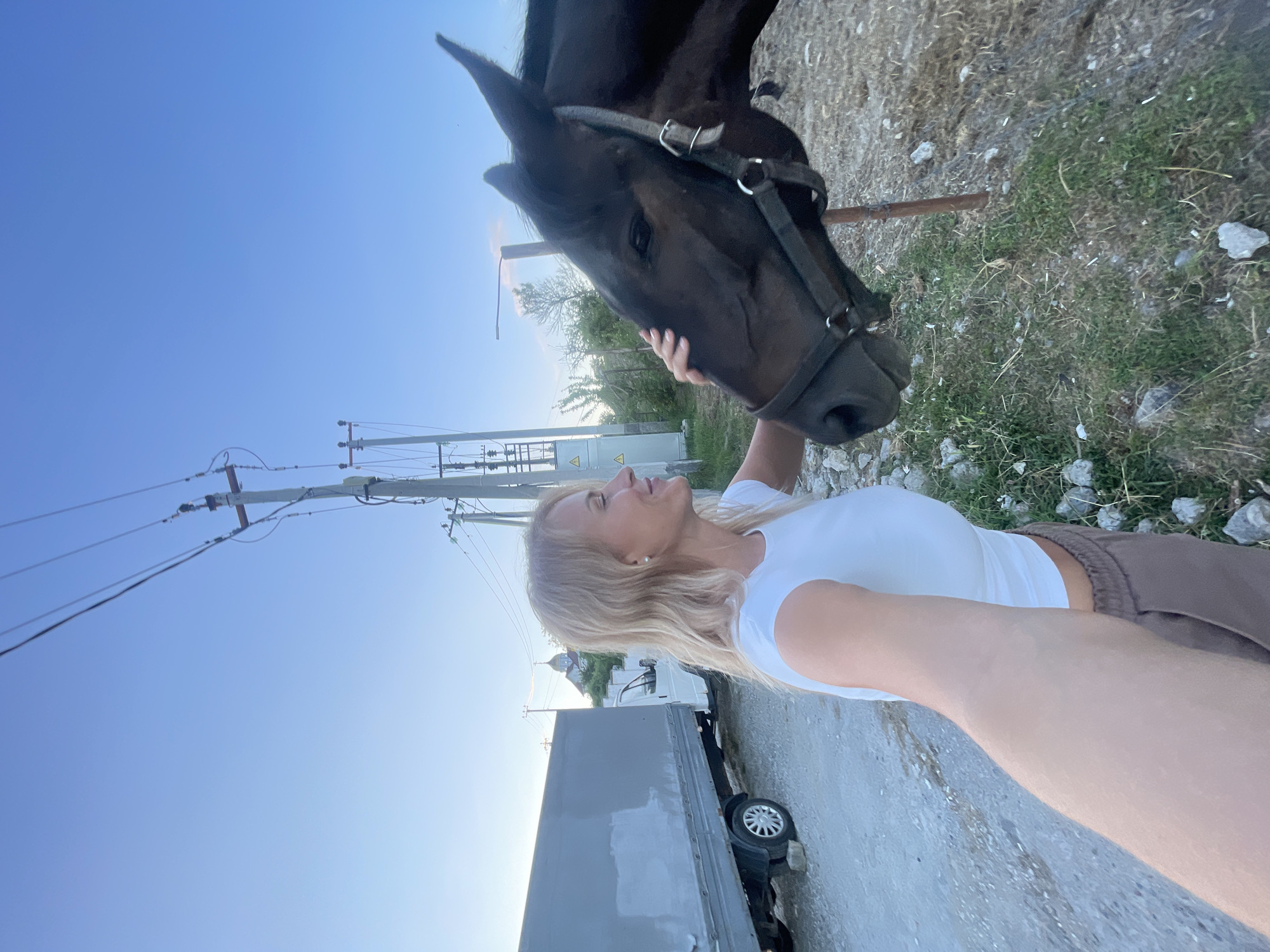 A blonde woman in a white crop top and brown sweatpants is standing outside, near a dark brown horse. She is holding her phone out, as if taking a selfie, and gently stroking the horse's head. The horse is wearing a bridle. In the background, there is a white truck with a gray tarped cargo area, power lines and poles, and some trees. The ground is covered in gravel and patchy grass. The sky is clear and blue. The scene appears to be in a rural or semi-rural area, possibly on the outskirts of Sochi, Russia.