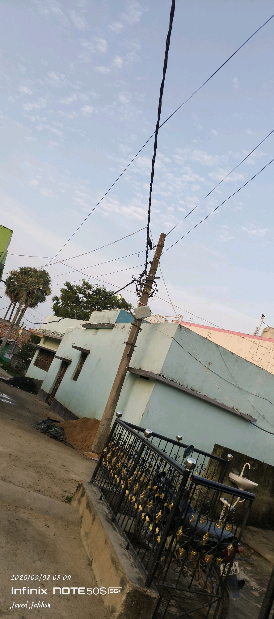 The image depicts an outdoor scene in what appears to be a residential area in Bahera, India, based on the provided text overlay. In the foreground, a black metal fence with decorative gold floral elements runs across the lower part of the frame. Behind the fence, a motorcycle is partially visible. To the left of the fence is a concrete pathway or sidewalk. Further back, a light blue and white house with a pink-trimmed roof is prominent. A wooden utility pole with multiple wires extending from it stands to the right of the house. Behind the house, palm trees and other vegetation are visible against a sky with scattered clouds. The ground is a mix of dirt and sand. The timestamp "2026/09/08 08:09" and the text "Infinix NOTE 50S SG+" and "Javed Jabbar" are watermarked on the bottom left of the image. There are no people visible in the frame.