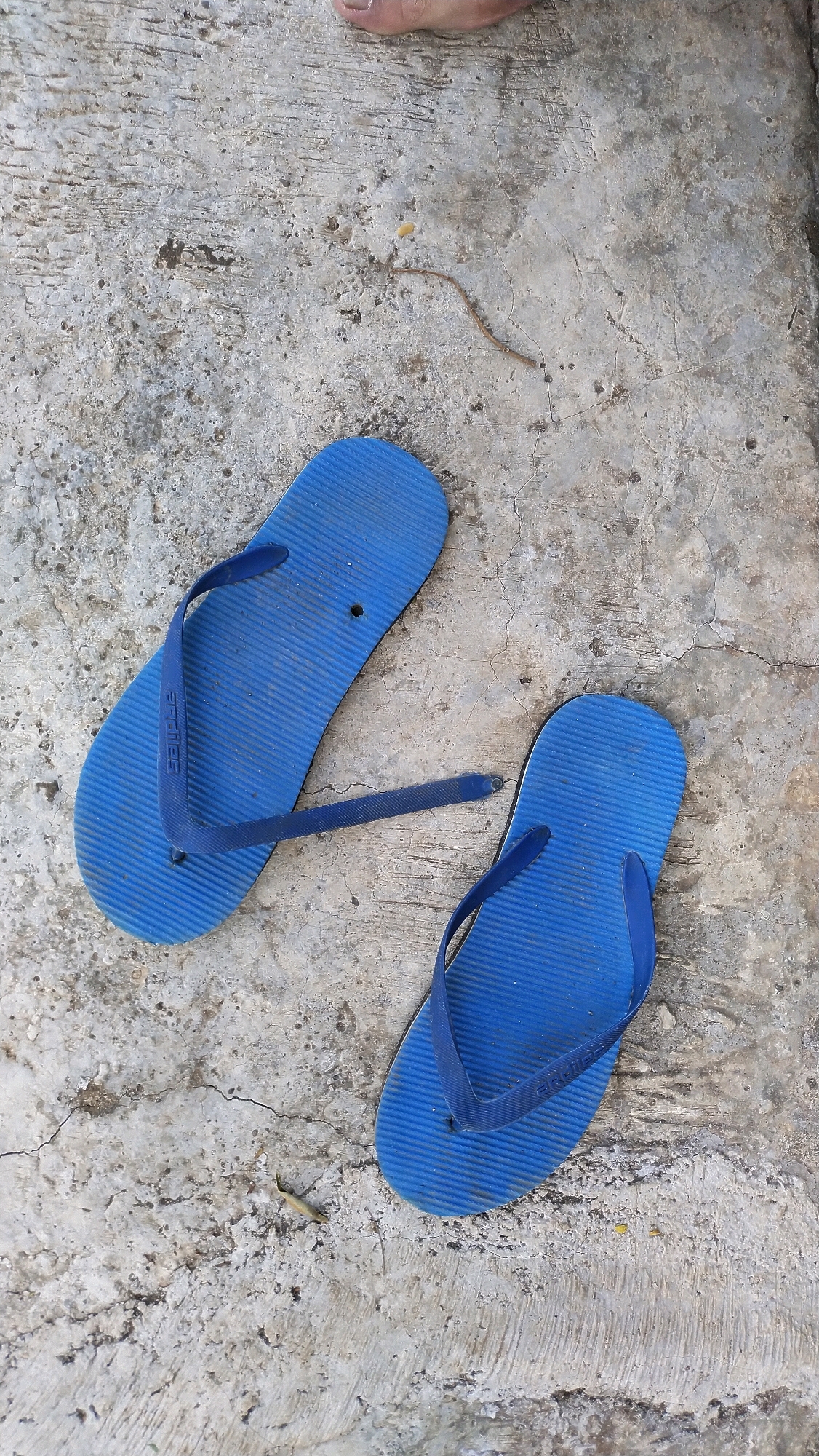 A pair of blue flip-flops rests on a cracked concrete surface. The flip-flops are positioned on their sides, with the soles facing upwards. One flip-flop is slightly above and to the left of the other. The concrete surface appears worn and weathered, with visible cracks and some dirt or debris scattered across it. A small, dried twig or plant stem lies on the concrete between the two flip-flops. The top edge of a human foot, with visible toes and fingernails, is partially visible in the upper portion of the image, suggesting the flip-flops may have recently been worn. This scene is located in Klungkung, Indonesia.