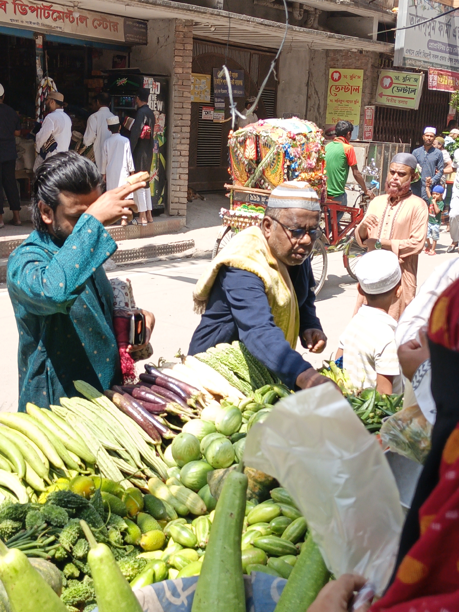 Here is a detailed description of the image:
**Setting**: The photo seems to have been taken at a street market or a busy urban area in what appears to be a South Asian country, possibly Bangladesh, given the Bengali script on the signs. The environment includes a mix of shops, street vendors, and pedestrians.
**Objects & Subjects**:
* **People**: Many people of different ages and attire are seen. Men mostly wear traditional clothing, with some wearing casual modern outfits. Children in traditional headwear are also present. One man in a teal-colored garment is holding a mobile phone to his face. Several individuals wear Islamic caps.
* **Vegetables**: There are many types of fresh vegetables displayed prominently in the foreground: gourds, eggplants, bitter gourds, and other leafy greens. A vendor is attending to a customer, placing vegetables into a plastic bag.
* **Cart**: A decorated hand-pulled cart with what looks like religious or cultural adornments stands out in the background.
* **Shops**: There are shops along the street with visible signage in Bengali, with text like "Departmental Store" and dental clinic advertisements.
**Actions & Interactions**:
* People are shopping, vendors are selling. There seems to be a general bustle of activity associated with a busy market.
* The man in teal holds a phone to his face, possibly taking a photo.
* The vendor is attending to a customer, placing vegetables into a plastic bag.
**Environment**:
* The urban environment suggests a densely populated area.
* Buildings are a mix of brick and concrete structures, some showing wear.
**Overall Impression**: The scene appears to be a typical day at a vibrant street market in an urban South Asian setting.