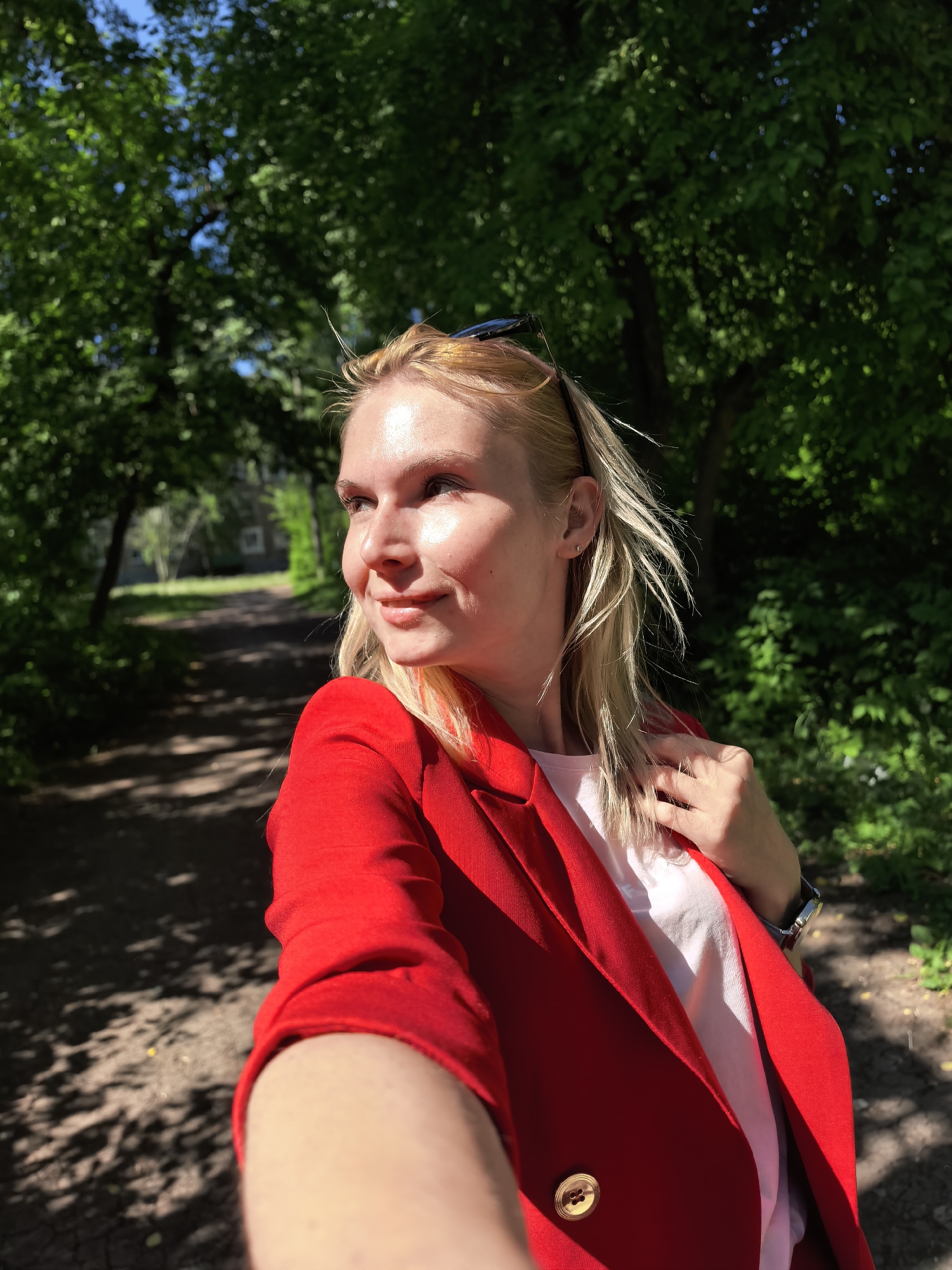 A blonde woman in a red blazer and white t-shirt is taking a selfie in a park. The woman is looking to the left. Behind her is a path through the park with trees on both sides. It is a sunny day and the trees are green. The woman has sunglasses on her head. The woman is wearing a watch on her left wrist. The location is Sterlitamak, Russia.