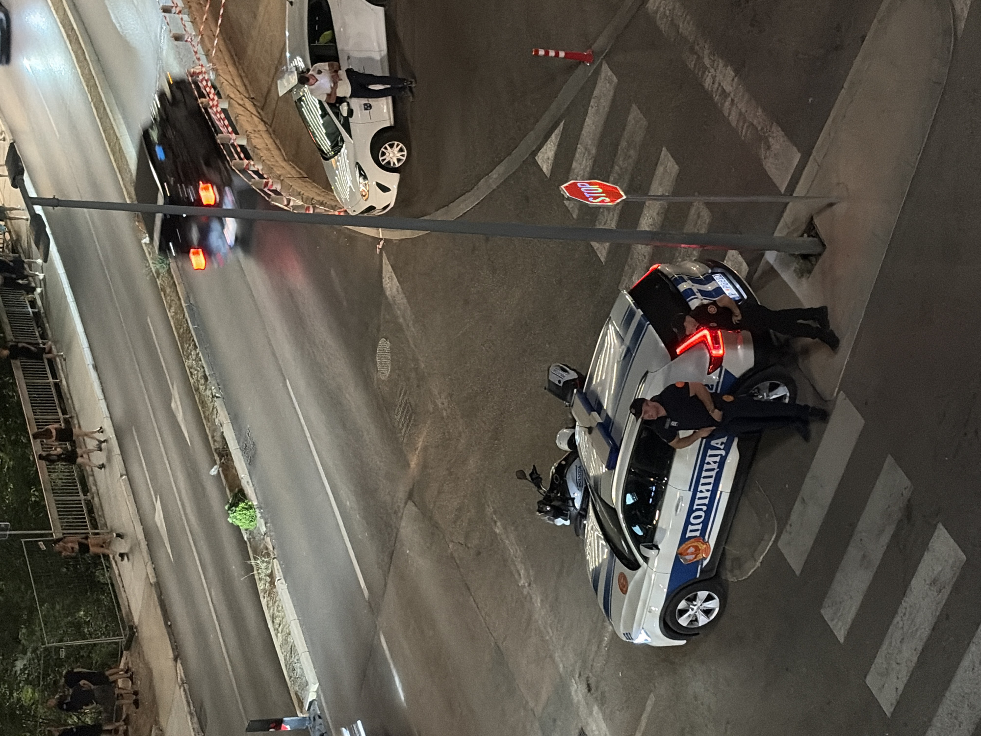At an intersection in Boreti, Montenegro, two police officers stand near a marked pedestrian crossing next to a police car. The police car, a white SUV with blue and white stripes and the word "ПОЛИЦИЈА" (POLICIJA) on its side, is parked on the right side of the road. A motorcycle is parked behind the police car. A stop sign is visible at the intersection. In the background, a dark-colored vehicle is blurred due to motion, suggesting it is moving at a high speed. Pedestrians are seen walking on a sidewalk in the upper left of the image. The scene appears to be taking place at night.