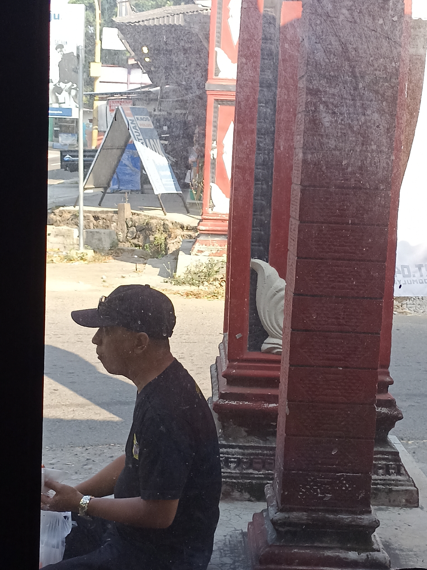 A medium shot captures a man wearing a black t-shirt and a black baseball cap, facing left in profile. He is seated on the ground with a white plastic bag to his left and a watch on his left wrist. The man appears to be in a shaded area, with sunlight casting shadows on the ground. Behind him, a red stone structure with decorative carvings and a white ornamental statue are visible. In the background, a street with an A-frame sign and a billboard can be seen. The overall scene suggests an outdoor urban environment. The image appears to be taken from a slightly elevated position, possibly from a vehicle. The location is identified as Pulisen, Indonesia.