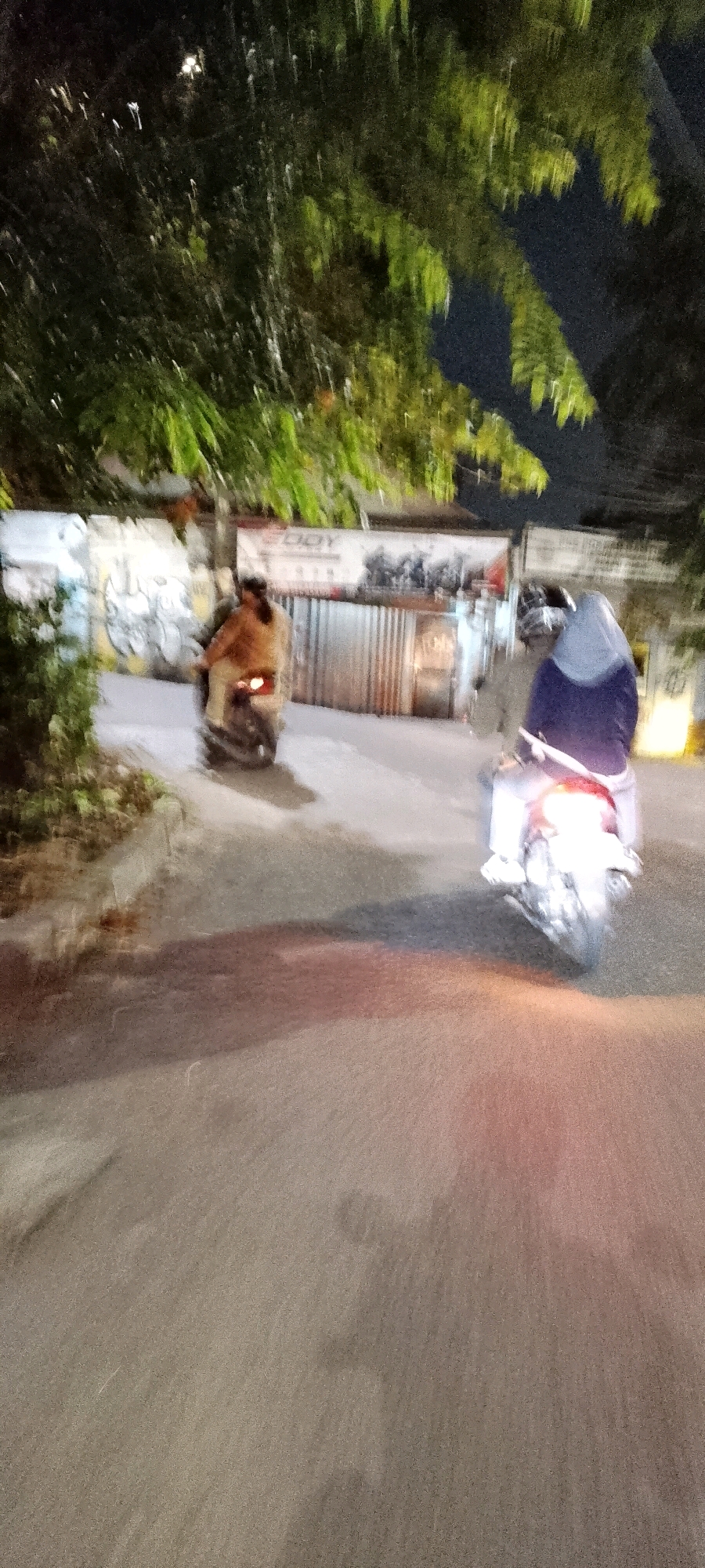 The image captures a blurred nighttime scene in Bekasi, Indonesia, showing two people riding motorcycles on a street. On the left, a person wearing a light brown jacket rides a motorcycle, with their face obscured. To the right, another individual, wearing a blue jacket and a headscarf, is on a motorcycle, their face also not clearly visible. Both motorcycles have their brake lights illuminated. The background features trees with green foliage against a dark sky, and a corrugated metal fence with some graffiti visible on a wall behind it. The street surface appears to be asphalt. The overall impression is one of motion blur, indicating the photograph was taken while the camera was moving.