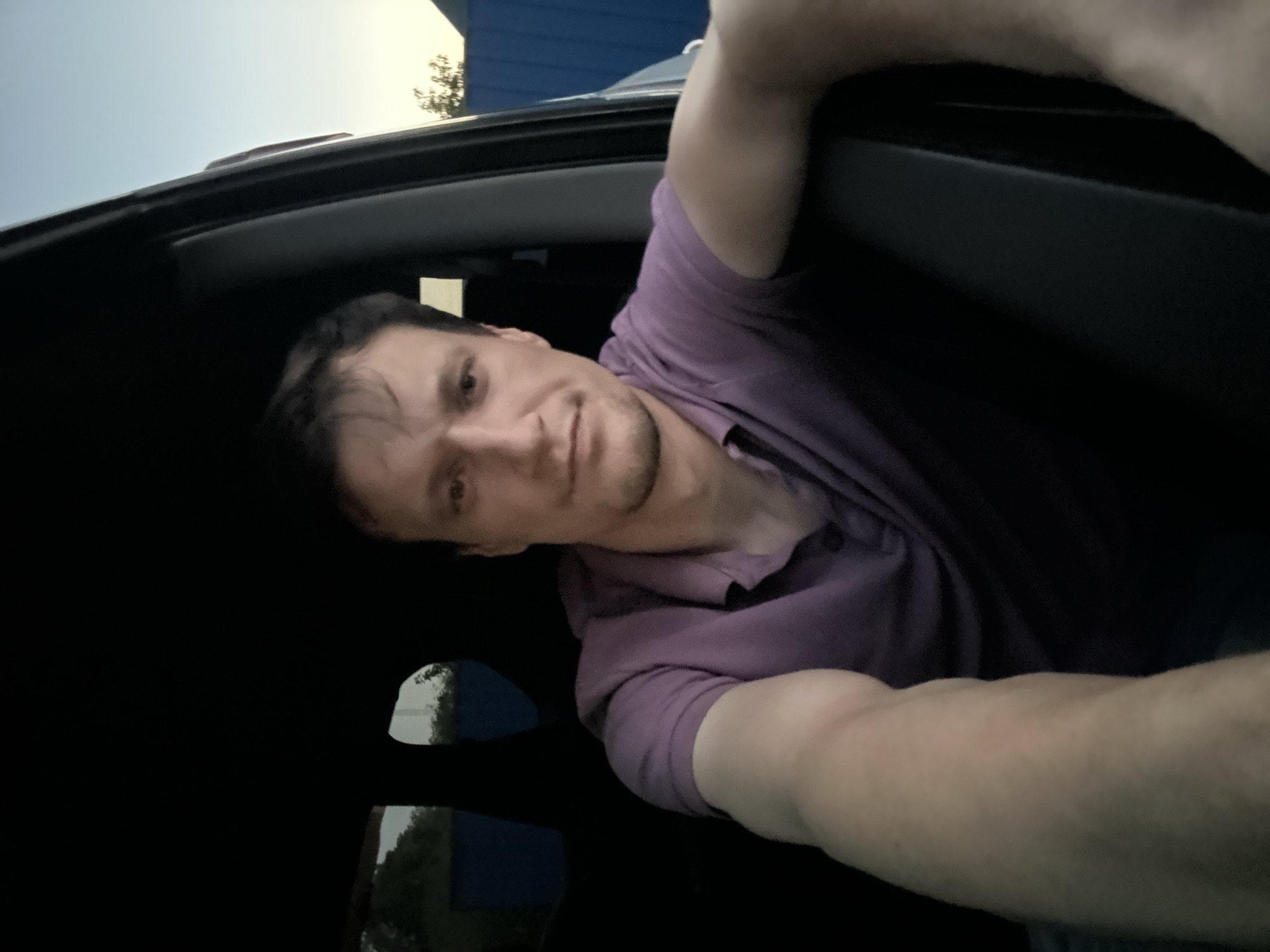 A man is sitting in a car, looking at the camera. He is wearing a purple polo shirt. The car appears to be parked, possibly in a residential area, as indicated by the fence visible in the background. The lighting suggests it is either dawn or dusk. The man's right arm is extended towards the camera, suggesting he is taking a selfie. The background shows a clear sky and some trees, along with a blue fence. The location is identified as Shatava, Ukraine.