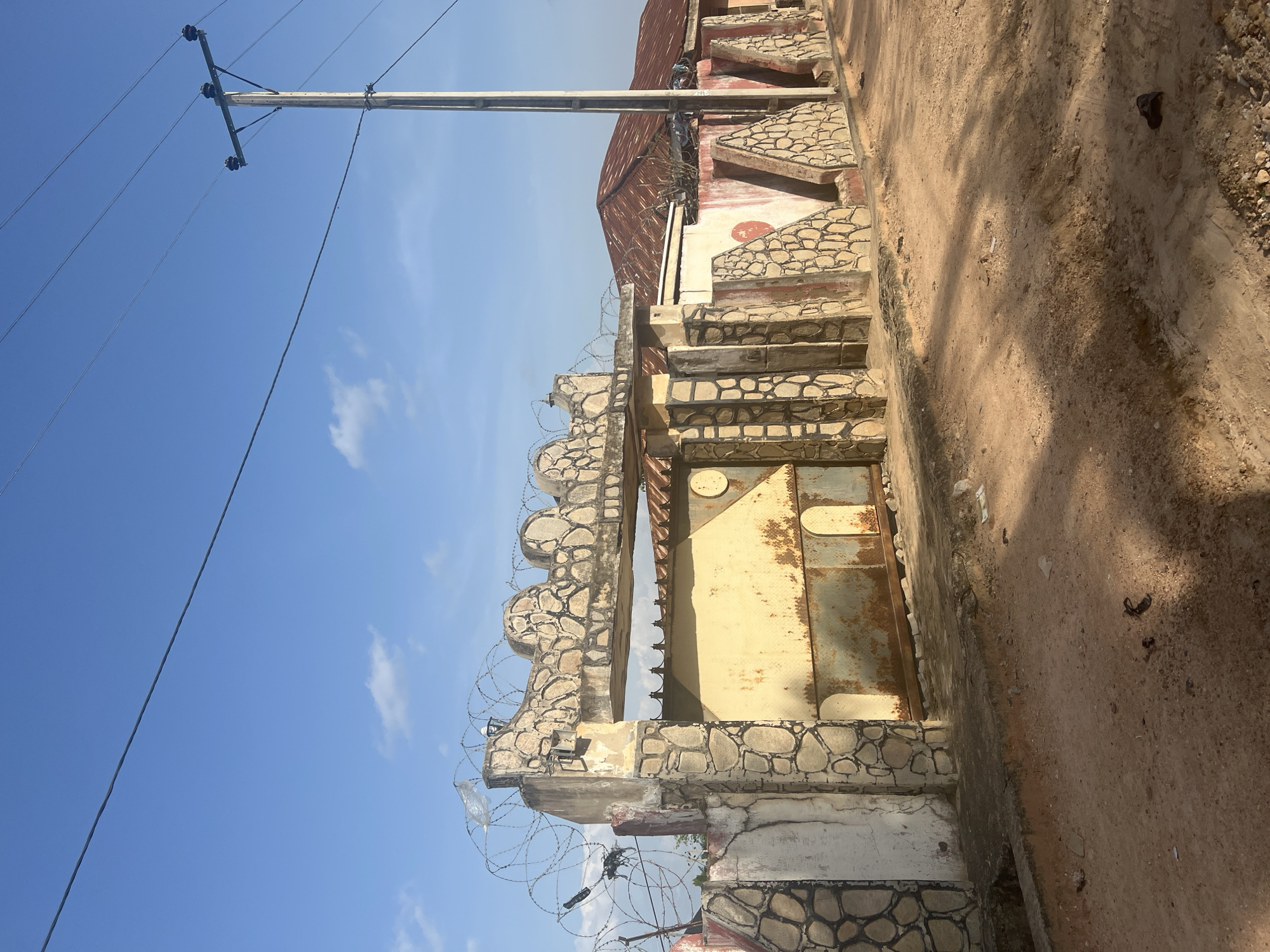 The image depicts an outdoor scene in Jalingo, Nigeria, featuring a property entrance. A large, weathered metal gate, painted yellow and showing signs of rust, is centrally positioned. The gate is flanked by a stone-decorated fence and pillars. The fence has a textured, light-colored surface adorned with darker, irregularly shaped stone patterns. Barbed wire is strung across the top of the gate structure.
Behind the gate, a portion of a building with a corrugated metal roof is visible. The building's exterior walls are partially painted white and red, with similar stone decorations to the front fence. A utility pole with power lines is prominent on the right side of the image, stretching across the blue sky. The ground in front of the entrance is a dirt road, characterized by loose soil, some scattered debris, and shadows cast by the sun. The overall impression is one of a functional, perhaps older, residential or commercial property in a dusty, sunlit environment.