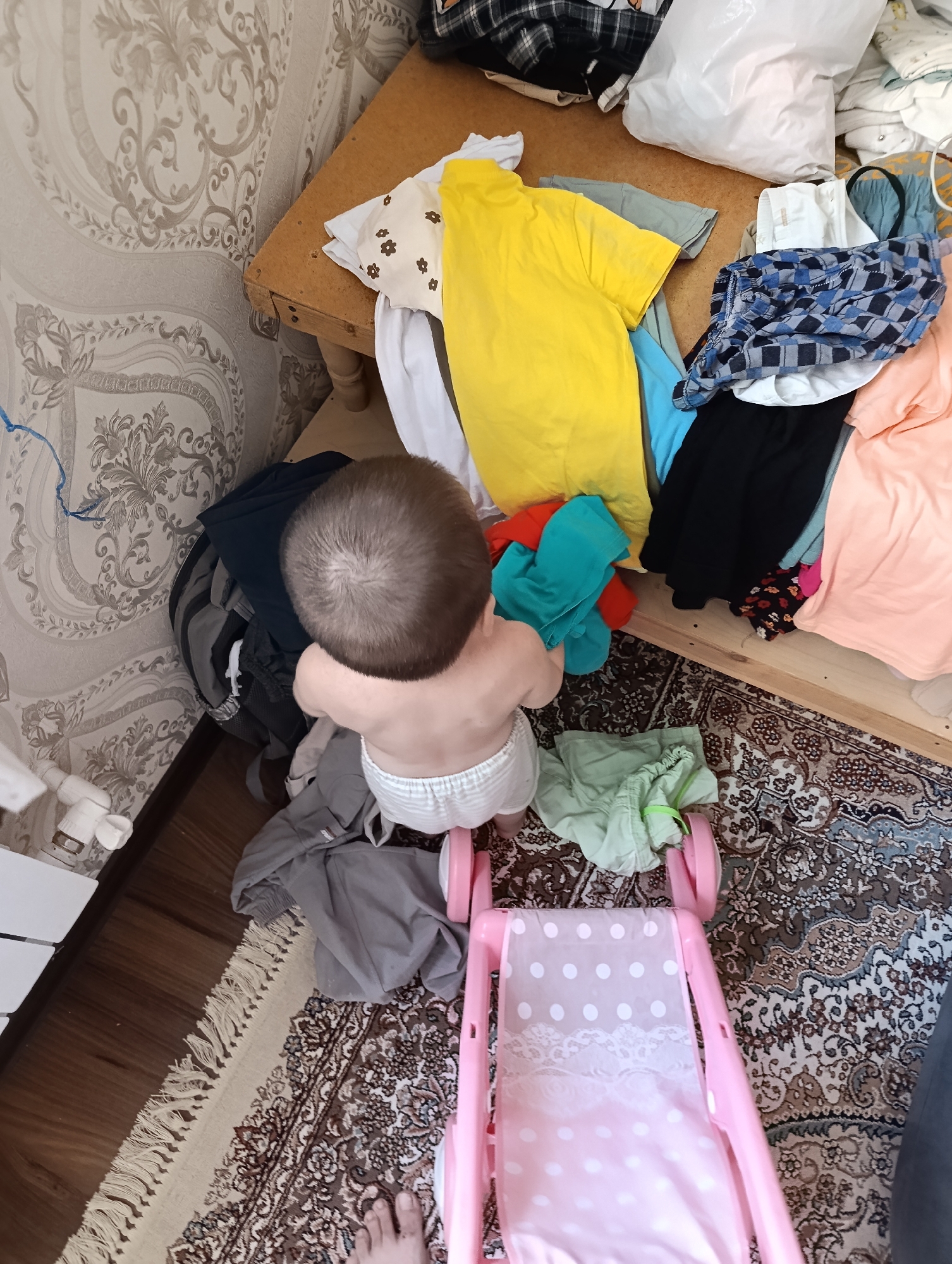 The image shows a messy room with clothes scattered around. A baby wearing a diaper is standing and reaching for some clothes. There's a wooden table or shelf with more clothes piled on it. A pink toy stroller is on the floor. The walls have patterned wallpaper, and a rug covers part of the floor. The scene appears to be an indoor residential setting. The location of Nukus, Uzbekistan, is not depicted or suggested by any elements within the image.