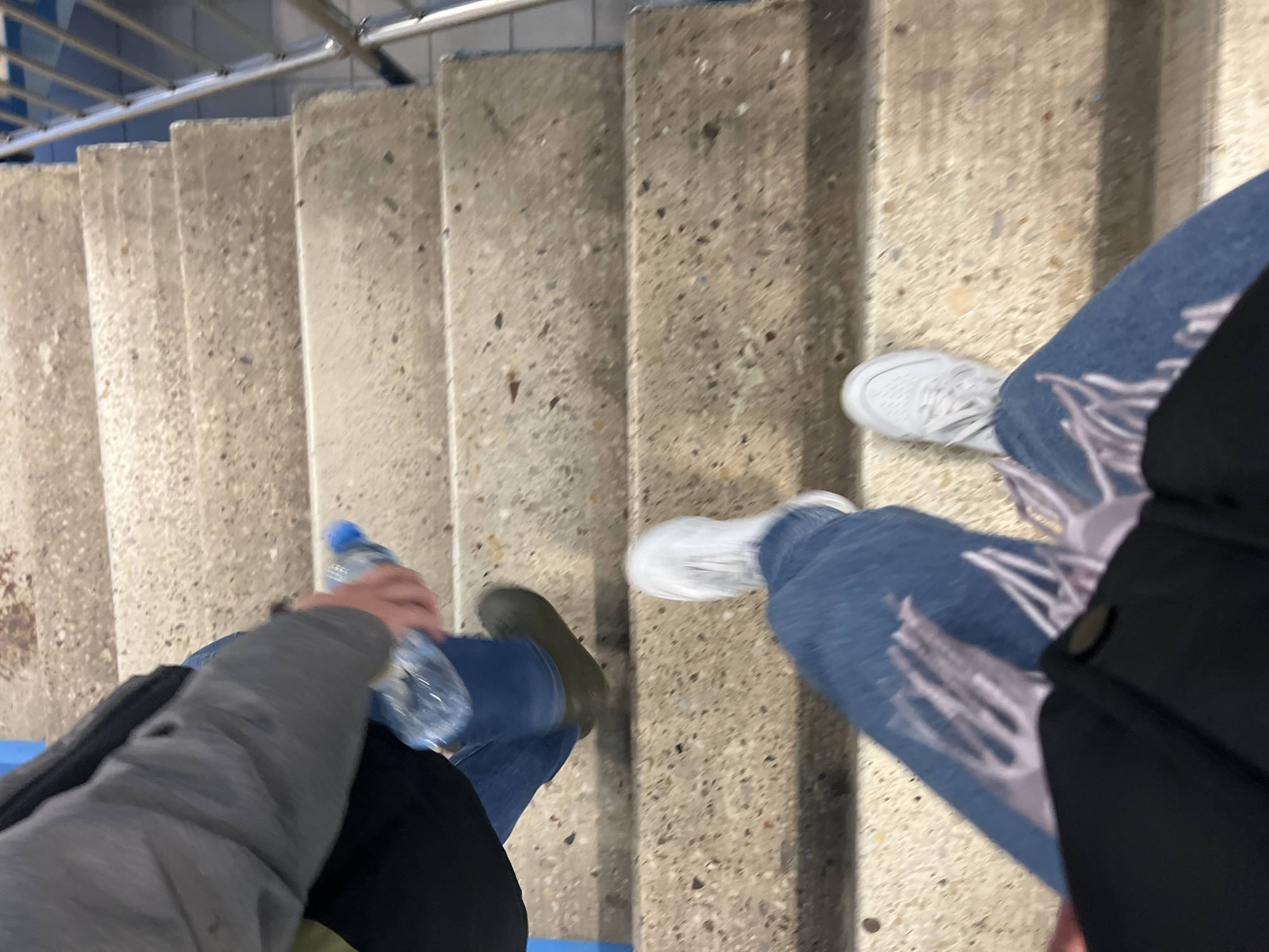 The image captures a downward-looking perspective of two individuals traversing a concrete staircase in what appears to be an indoor or covered public setting in Khimki, Russia.
On the left side of the frame, one person is seen wearing blue jeans and a grey long-sleeved garment, possibly a hoodie or jacket. Their right hand holds a clear plastic water bottle with a blue cap. One foot, wearing a dark, possibly olive green, shoe or boot, is visible on a lower step. A black bag or jacket is draped over their left side, partially obscuring the other foot.
On the right, another person is visible, wearing light blue jeans featuring prominent white or light-colored graphic designs, which appear to be stylized hands or abstract patterns. They are wearing white sneakers. A black jacket or coat is visible on their right side.
The staircase itself is constructed from light grey concrete with a speckled aggregate texture. A metallic handrail with several horizontal bars runs along the left side of the steps, with a backdrop of blue tiles or a painted wall. Motion blur on the feet and legs of both individuals suggests they are in the process of descending the stairs. No visible text or specific time of day cues are present in the image.