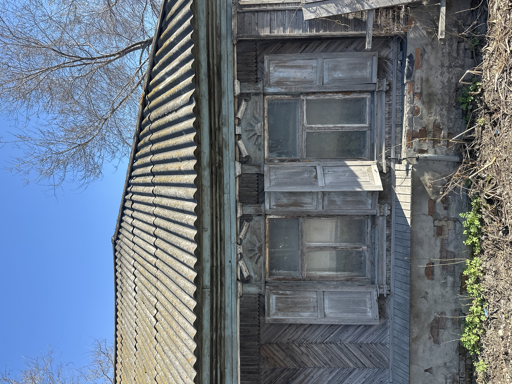 The image, rotated 90 degrees counter-clockwise, depicts a close-up, upward-angled view of a weathered wooden house against a clear blue sky in Nogornyi, Russia. The corrugated asbestos cement roof, with its distinct ridges and shadows, dominates the upper portion of the frame, showing signs of age and discoloration.
Below the roofline, the house's upper facade is constructed from dark, weathered wooden siding, featuring both vertical planks and an intricate herringbone pattern. Two grimy windows with faded wooden frames and multiple panes are visible, flanked by decorative wooden surrounds. These surrounds include carved elements, most notably a stylized sun-like motif positioned between the windows. The wooden window shutters appear aged and unpainted, with one seemingly partially open.
To the right of the wooden facade, a section of a concrete or stucco wall shows significant deterioration, with patches revealing underlying red bricks. A narrow pipe or conduit runs along this exposed wall. Along the far right edge, a mix of dried, matted vegetation and newly sprouted small green plants suggests an overgrown area near the ground level. Above the roof on the left, the bare branches of a deciduous tree are silhouetted against the bright, cloudless blue sky, indicating a late winter or early spring setting. The scene is illuminated by clear, direct sunlight, suggesting daytime. No activity is taking place, and there is no visible text.