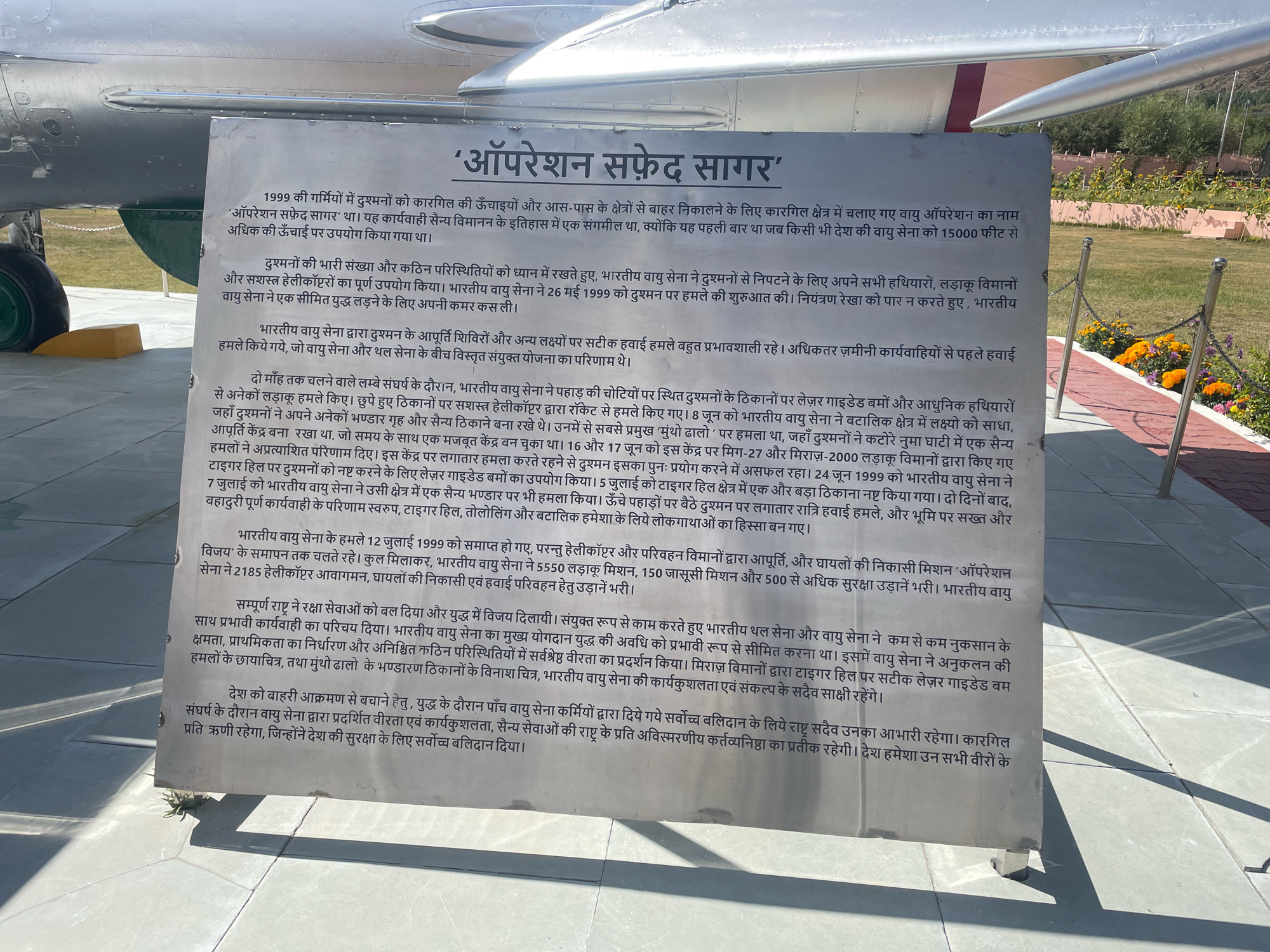 The image shows a plaque in front of a military aircraft. The plaque is titled "'Operation Safed Sagar'" and is written in Hindi. The text describes the role of the Indian Air Force in the Kargil War of 1999. The aircraft appears to be an older model, possibly a fighter jet, with visible landing gear and part of the fuselage. The background shows a landscaped area with flowers and a chain-link fence, suggesting it is an outdoor exhibit or memorial. The location is identified as Bhimbat, India, in the description provided with the image. The lighting suggests it is daytime, and the overall scene is one of historical commemoration.