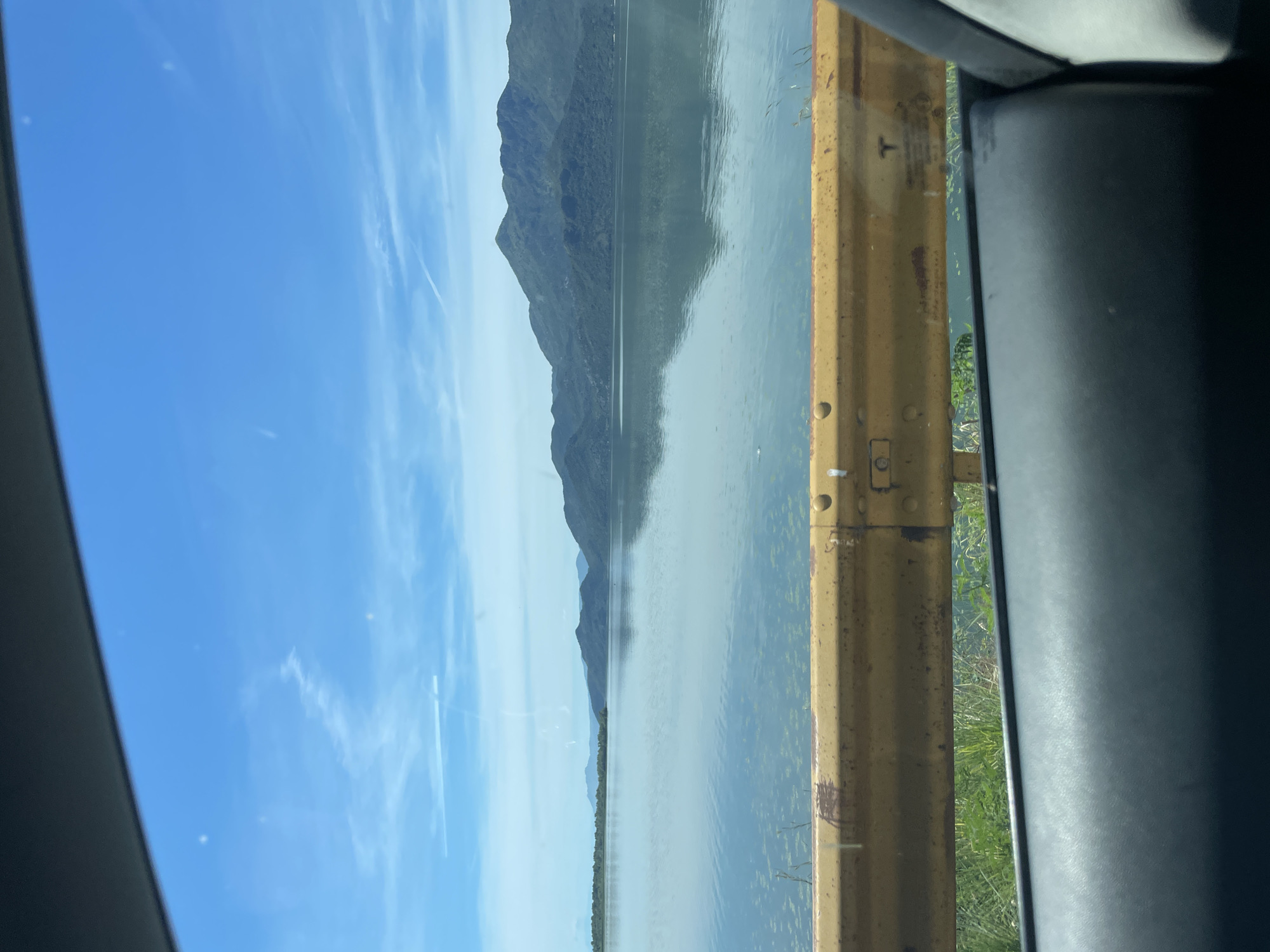 A wide, calm body of water, likely a lake, reflects a hazy sky with scattered white clouds. The water's surface is mostly smooth, with some patches of what appear to be aquatic vegetation. In the background, a range of dark, undulating hills or mountains stretches across the horizon, their slopes covered in dense foliage. In the foreground, a weathered, bright yellow guardrail runs horizontally across the frame. Below the guardrail, lush green grass and plants are visible. The bottom of the image shows a dark, textured surface, likely part of a vehicle's interior, suggesting the photo was taken from inside a car. The scene appears to be the lake shore near the village of Vranjina, Montenegro, known for its proximity to Skadar Lake, a large freshwater lake bordered by Montenegro and Albania.