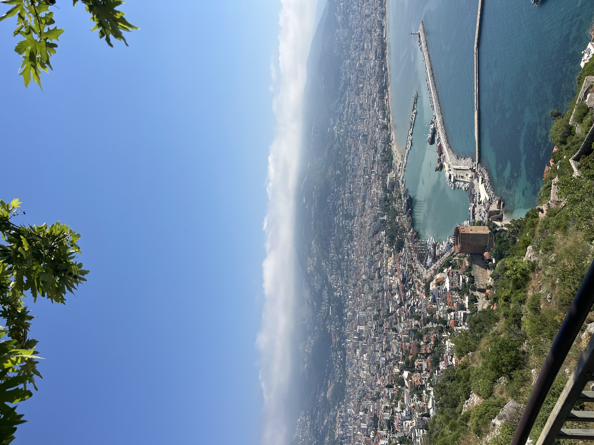A wide, high-angle panoramic photograph captures the extensive coastal city of Alanya, Turkey, under a clear blue sky. The image is taken from an elevated viewpoint, likely a hilltop or castle, offering a sweeping vista over the urban landscape, harbor, and sea.
In the upper-left and upper-right corners, vibrant green leaves from a tree branch partially frame the sunny sky, indicating the photographer's position beneath foliage. The sky above is a brilliant blue, with a soft band of white, hazy clouds hugging the distant mountain range that backs the city.
The city of Alanya sprawls across the center and left of the frame, showcasing a dense cluster of buildings, residential areas, and commercial structures extending along the coastline and climbing the lower slopes of the mountains. Towards the right side of the image, the expansive harbor is a prominent feature, protected by long, concrete breakwaters. Numerous boats, including yachts and smaller vessels, are moored within its calm turquoise waters. The deeper blue of the open Mediterranean Sea stretches beyond the harbor.
A distinctive red-brown, octagonal stone tower, the Kızıl Kule (Red Tower), stands out on the coastline near the harbor entrance, a historical landmark against the modern city. The immediate foreground on the lower-right reveals a metal railing, suggesting the vantage point is a public overlook or a secure viewing area.
The scene is bathed in bright daylight, indicating mid-day sun. The overall impression is one of a bustling coastal town enjoying good weather, with no specific human activity visible due to the distance. No discernible text is present in the image.