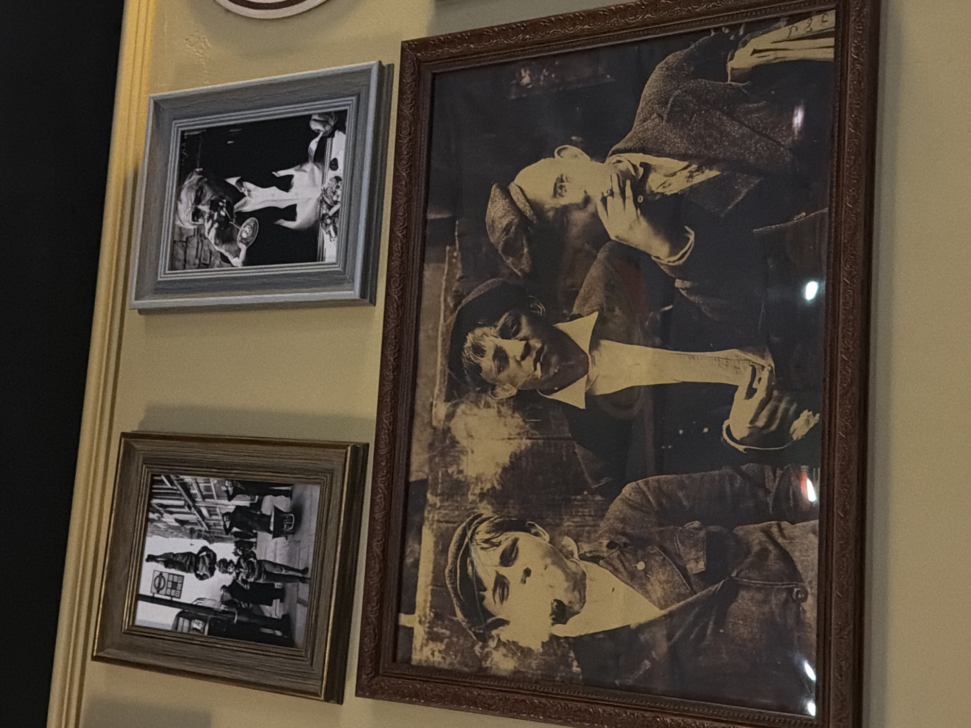 A wall features three framed photographs. The largest photograph, framed in dark brown, depicts three men in sepia tones, wearing caps and suits. The middle man wears a white collared shirt. The upper man has a hand to his face. To the left are two smaller framed black and white photographs. The top photograph, framed in gray, features a person holding a glass, seemingly pouring liquid into someone's mouth. The bottom photograph, framed in brown, captures a street scene with people, a scooter, and a sign featuring a red circle with a blue bar through the middle. The wall itself is painted a light beige. The scene is in Rostov-on-Don, Russia.