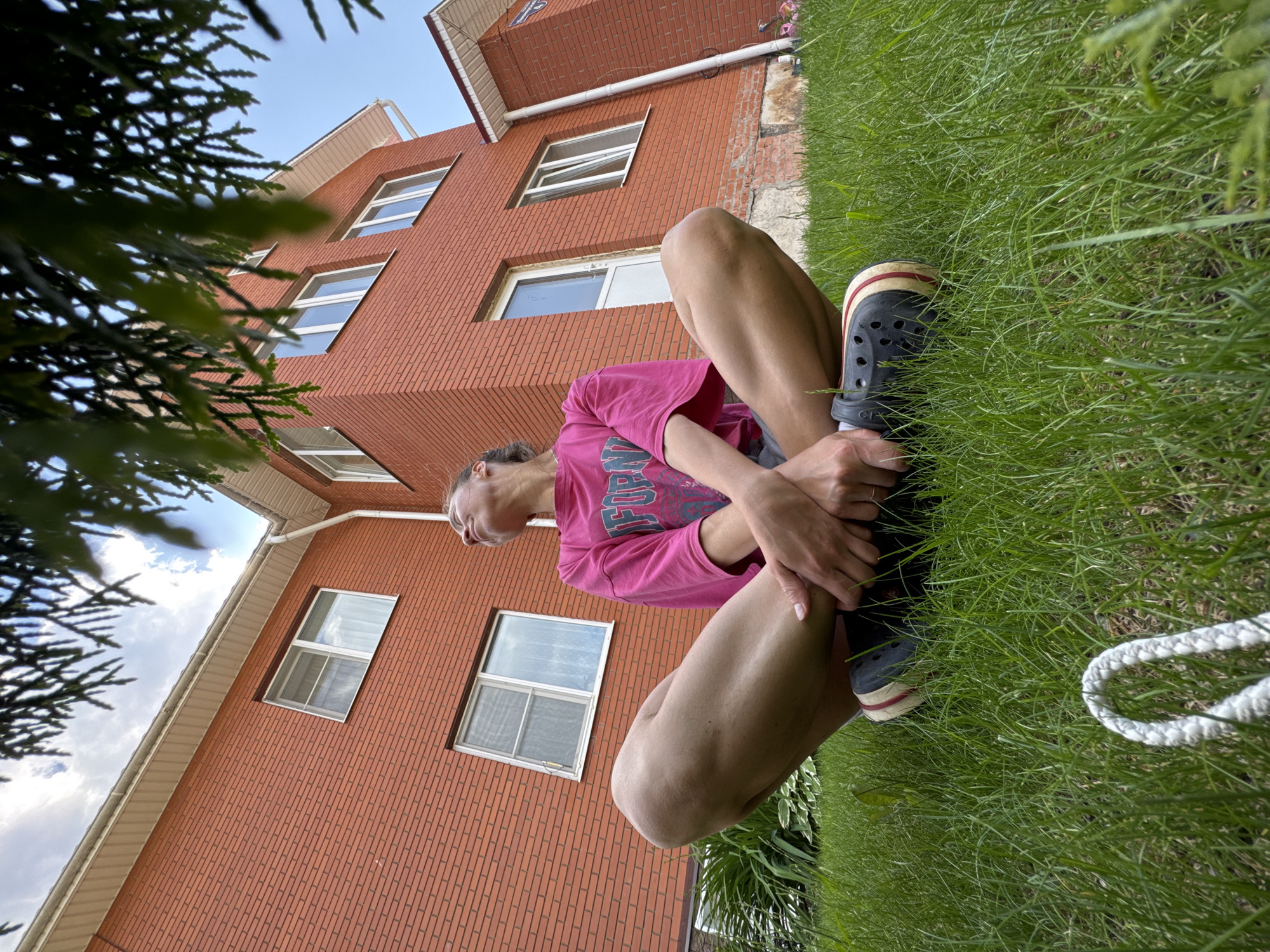 In Прикамский, Russia, a woman sits cross-legged on a vibrantly green, well-maintained lawn in front of a two-story brick house. The house, constructed with red bricks, features white-trimmed windows and a light-colored roofline. A dark green tree branches partially obscure the upper portion of the image, framing the scene from above. The woman is wearing a bright pink t-shirt with graphic text and dark grey shorts. Her legs are crossed, with her hands resting on her shins, and she is looking upwards and to her left. She is wearing dark grey clogs with red and white trim. In the foreground of the lawn, a portion of a white rope is visible. The sky above is partly cloudy.