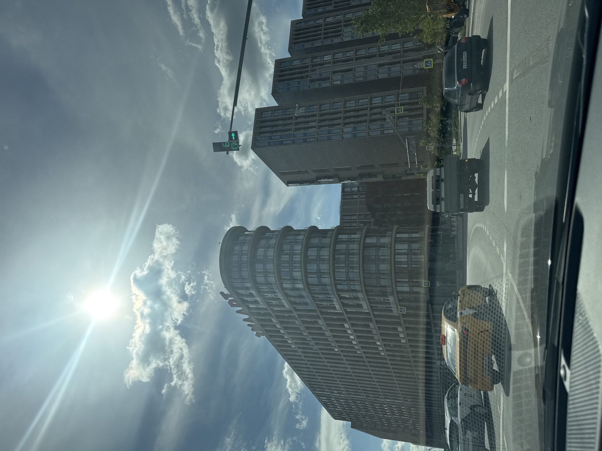 The image shows a street scene in Moscow, Russia, on a sunny day with some clouds. The foreground features the interior of a vehicle, with a dashboard and windshield visible. Several cars are on the road, including a yellow taxi and a dark sedan. In the background, modern buildings with many windows are prominent. The largest building, on the left, is curved and features the word "SMART" in red lettering at the top. A traffic light with a green arrow is visible above the road. The sun is bright and shining through the clouds, creating lens flare.