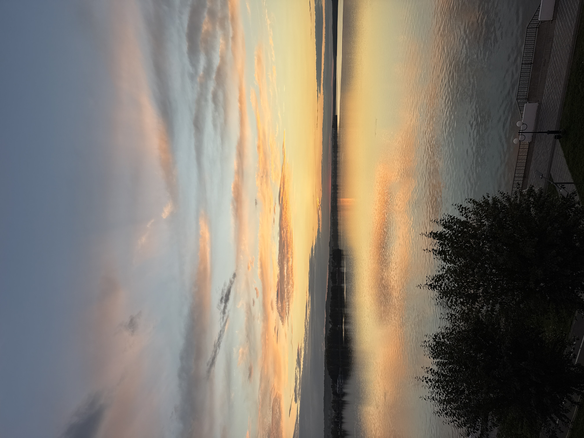 The image depicts a tranquil sunset over the Votkinsk Reservoir in Votkinsk, Russia. The sky is filled with soft hues of orange, pink, and blue, with wispy clouds reflecting on the calm water. In the distance, the silhouette of the city's shoreline is visible, dotted with trees and buildings. The foreground features lush green trees and a paved promenade with a decorative lamppost. The scene suggests a peaceful evening setting.