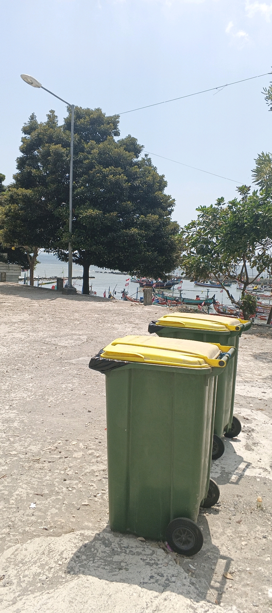 In Kandangsemangkon, Indonesia, on a sunny day, two green trash bins with bright yellow lids are positioned on a paved area near a body of water. The bins are slightly angled, with the one on the left closer to the foreground. They both have black plastic liners peeking out from under the lids. Beyond the bins, the scene opens up to a waterfront with numerous small fishing boats docked. Lush green trees frame the view of the boats and the calm, blue water. A single lamppost stands tall on the left side of the image, its pole extending upwards against a clear sky. The ground is a mix of paved sections and dirt, with scattered debris.