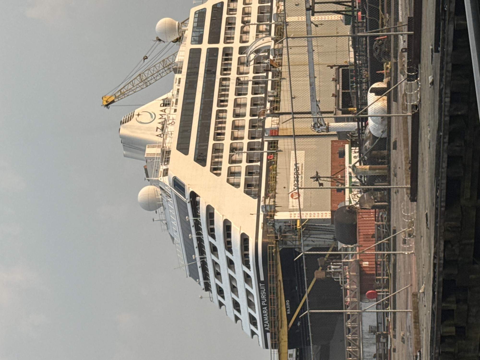 A large cruise ship named "AZAMARA PURSUIT" is docked at a shipyard in North Vancouver, Canada. The ship is white with multiple decks of windows and a distinctive funnel with a logo. A yellow crane is positioned over the ship. In the foreground, a chain-link fence separates the viewer from the shipyard. Various industrial equipment, shipping containers, and debris are visible on the dock behind the fence. The sky is overcast and appears to be early morning or late afternoon due to the soft lighting.