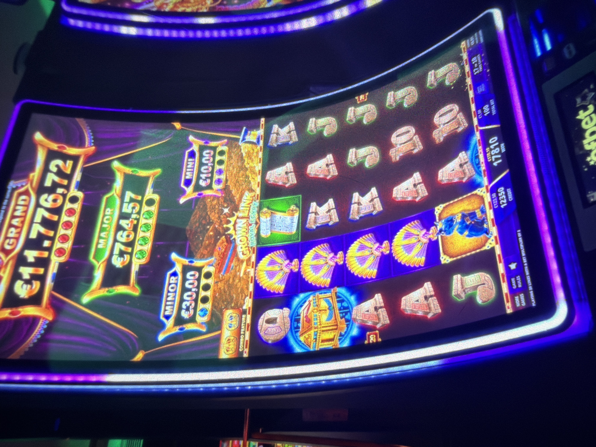 The image displays a close-up, eye-level view of a brightly lit, modern slot machine inside a casino. The machine's screen is the primary focus, showcasing a vibrant, animated display with gold and purple hues, featuring symbols such as stylized fans, ancient Egyptian-themed objects, and card game icons (A, K, J, 10). Prominently displayed are jackpot amounts: "GRAND €11.776,72", "MAJOR €764,57", "MINOR €30,00", and "MINI €10,00". Below the jackpot displays, the reel area shows a sequence of icons, including a golden chest with a blue aura. The bottom section of the screen displays credit, win, and bet information.
The slot machine itself is sleek and curved, with prominent blue and purple LED lighting illuminating its edges. The surrounding environment is dark, with only hints of other slot machines visible on the right and a dark curtain-like material in the background. The floor is not visible.
The scene is devoid of any people. The image appears to be taken in the city of Sofia, Bulgaria, within an establishment that offers gambling, likely a casino or a gaming hall.