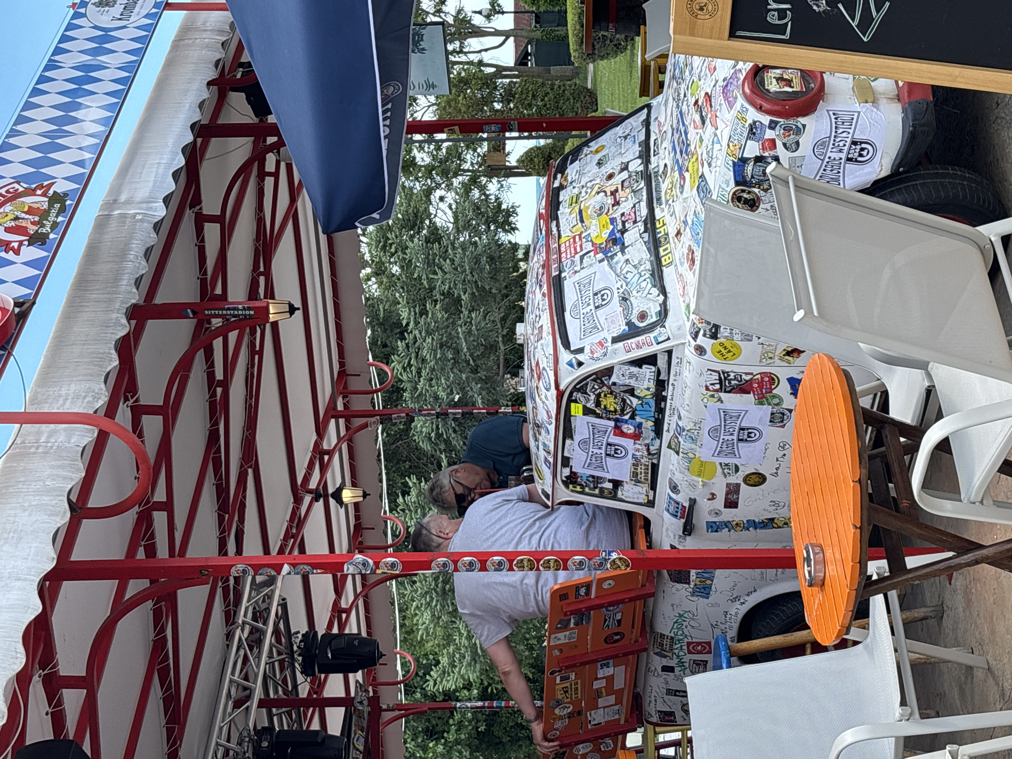 The image depicts an outdoor seating area with a unique, heavily stickered car serving as a central decorative element. Two men are seated inside the car, facing away from the camera, engaged in conversation. The car is white and covered with a multitude of colorful stickers, including branding and slogans. The seating area features an orange circular table with an ashtray, surrounded by several white metal chairs. In the background, lush green trees suggest a park-like setting. A red and white striped canopy extends over the scene, supported by red metal poles. The overall atmosphere is casual and likely a spot for outdoor dining or recreation. This scene is located in Varna, Bulgaria, at a location called "Brigade Weststadt", as indicated by stickers on the car and the overall theme of the establishment.