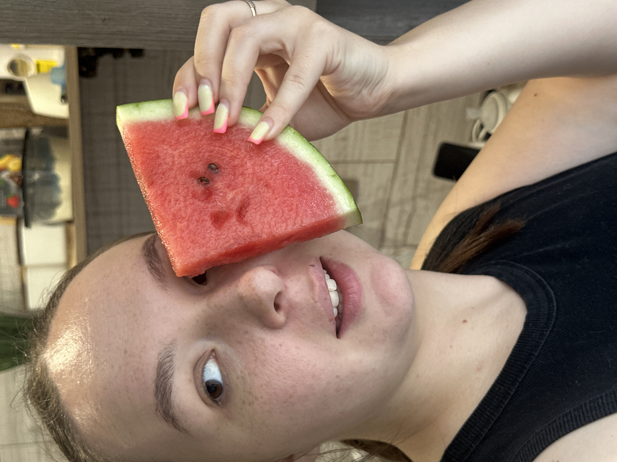 The image features a young person holding a slice of watermelon in front of their face, obscuring their eyes. The watermelon slice is a vibrant red with visible seeds and a green rind. The person has freckles and is wearing a black top. Their hand, adorned with a ring and light-colored nails with pink tips, is holding the watermelon. The background is blurred, showing wooden flooring and shelves with various objects, suggesting an indoor setting, possibly a kitchen or living area. The lighting appears natural, indicating it might be daytime. The overall mood is playful and lighthearted. There is no visible text in the image.