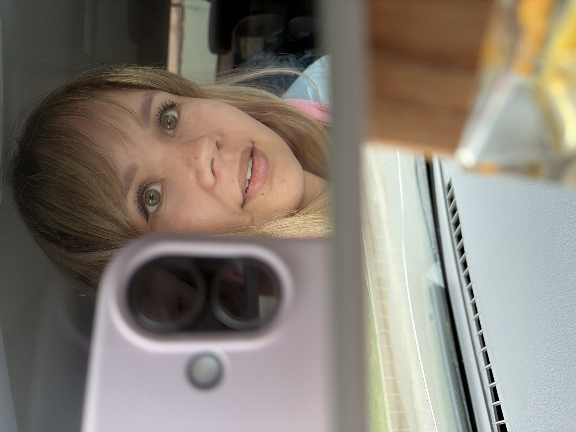 The image shows a selfie taken by a person with blonde hair and green eyes. The person is holding a light purple smartphone with two camera lenses visible. The reflection of the person is visible in the lenses. Behind the person, a car window with visible road and landscape outside. The location is unspecified.
