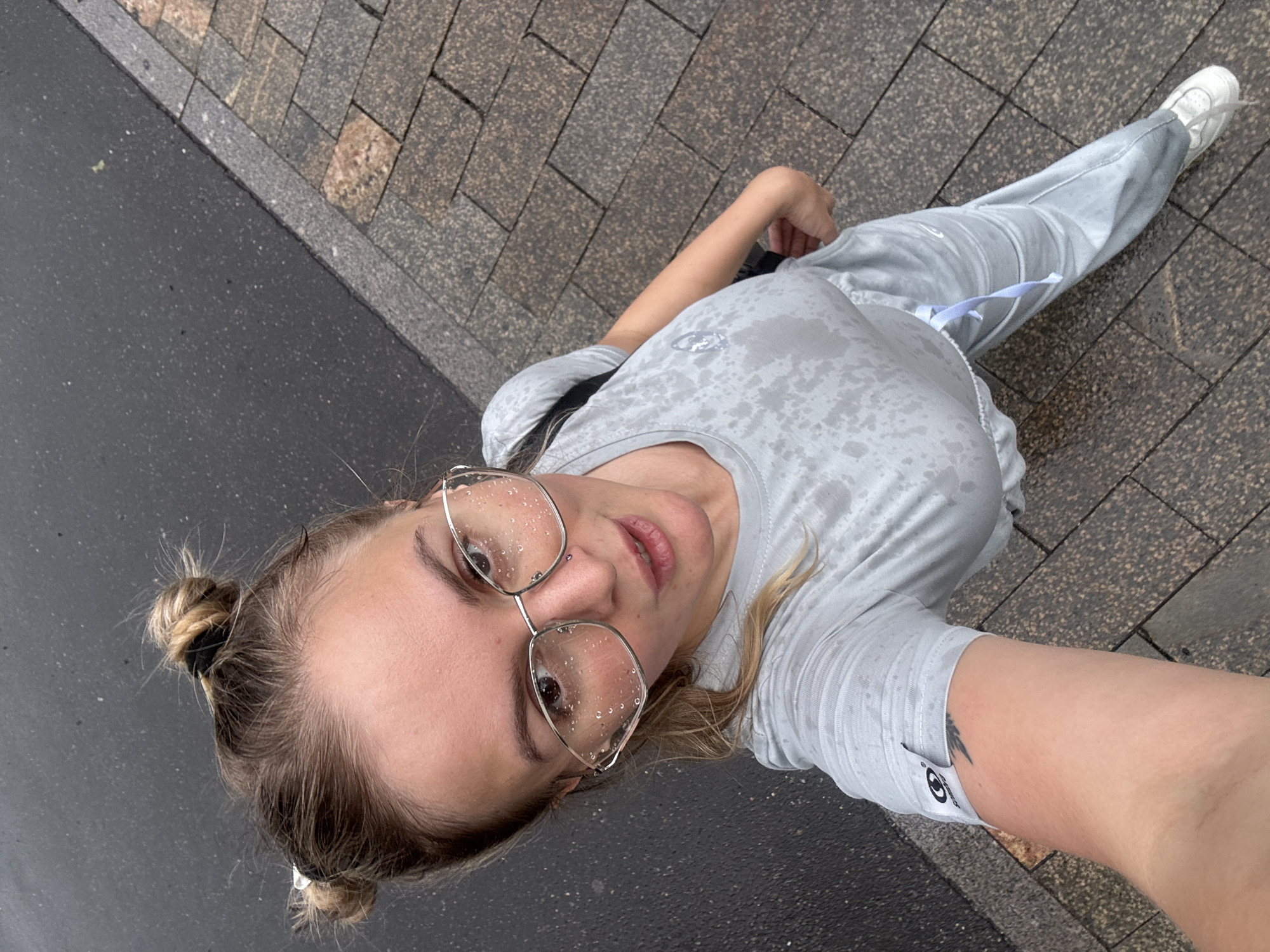 A woman stands on a wet sidewalk, wearing a light grey athletic outfit and clear-rimmed glasses. Water droplets are visible on her glasses and shirt. Her hair is styled in two buns on top of her head. The ground appears to be wet asphalt next to a brick sidewalk.
