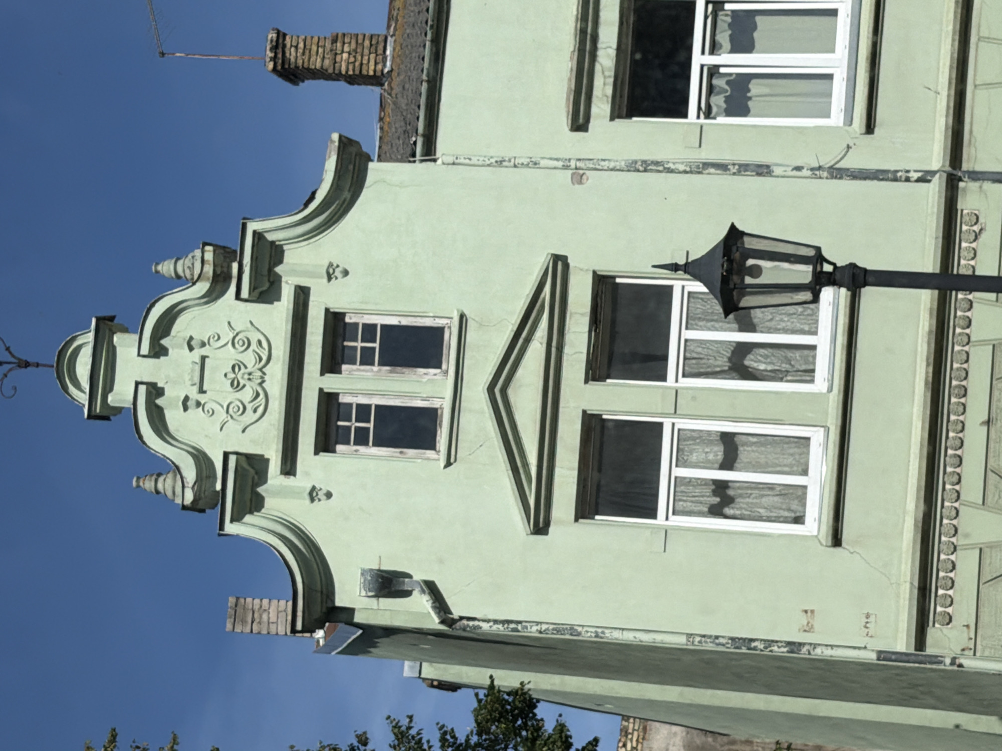 The image shows a pale green building with a stepped gable roof, characteristic of Baroque architecture, under a clear blue sky. The building has several windows; the upper two are narrower and divided into panes, while the lower two are larger and also divided. A decorative frieze with circular motifs runs along the lower part of the visible facade. A black, ornate lamppost stands in front of the building, slightly to the right of the center. To the right, a chimney made of red brick rises from the roofline, topped with an antenna. To the left, a section of another building, partially obscured by trees, is visible. The facade exhibits some minor cracks and signs of wear. This building is located on Lielais prospekts, Ventspils, Latvia, and is identified as house number 30.