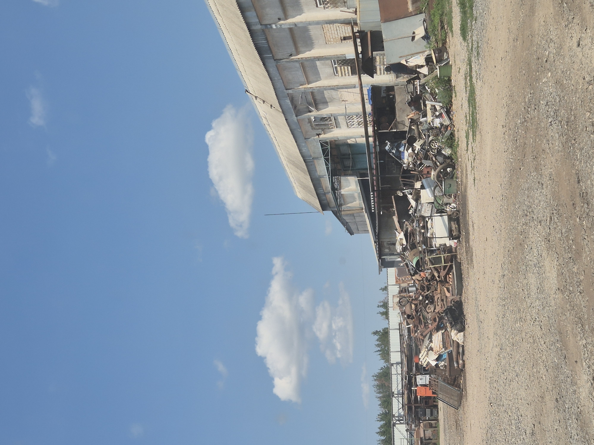 This image depicts an outdoor industrial or scrapyard scene located in Pidhaitsi, Ukraine. In the foreground, there is a large pile of scrap metal and various discarded items, including what appears to be vehicle parts, machinery components, and metal sheeting. The scrap is predominantly brown and rusty, with some lighter colored materials interspersed.
Behind the scrap pile, a long, industrial-style building with a corrugated metal roof and concrete walls is visible. The building appears weathered and somewhat dilapidated, with multiple windows, some of which are boarded up or have complex grates. A covered awning extends from the front of the building over a section of the scrap.
A person, dressed in dark clothing, is standing to the right of the building, near a large piece of metal. The ground throughout the scene is covered in gravel and dirt, with some patches of green grass growing at the edges. In the background, a line of trees forms a backdrop, beyond which a clear blue sky with scattered white clouds is visible. The overall impression is one of an active, albeit somewhat disorganized, scrap processing or storage facility.