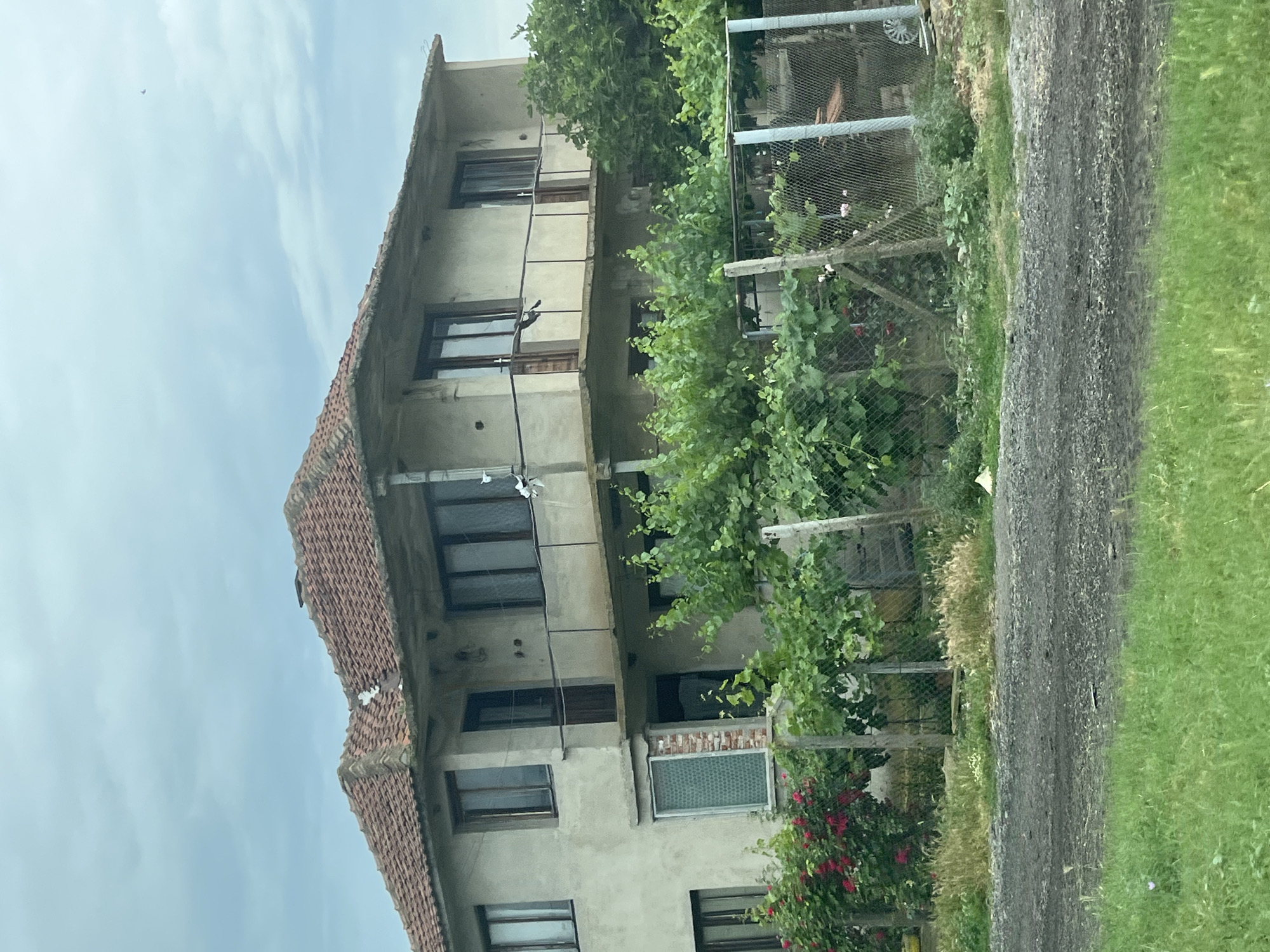 The image shows a two-story concrete house with a red-tiled roof in an exterior setting. The house is weathered and appears to be in a state of disrepair, with cracked concrete and missing roof tiles. There are several windows and doors on both floors, some with wooden frames. A balcony runs along the second floor, supported by concrete pillars. The house is surrounded by greenery, including trees, bushes, and flowering plants, with a chain-link fence separating the property from the foreground. A dirt road is visible in front of the fence, and beyond that, a grassy area. The sky is overcast with clouds. No people are visible in the image. The scene suggests a rural or semi-rural location, likely in a somewhat neglected state.