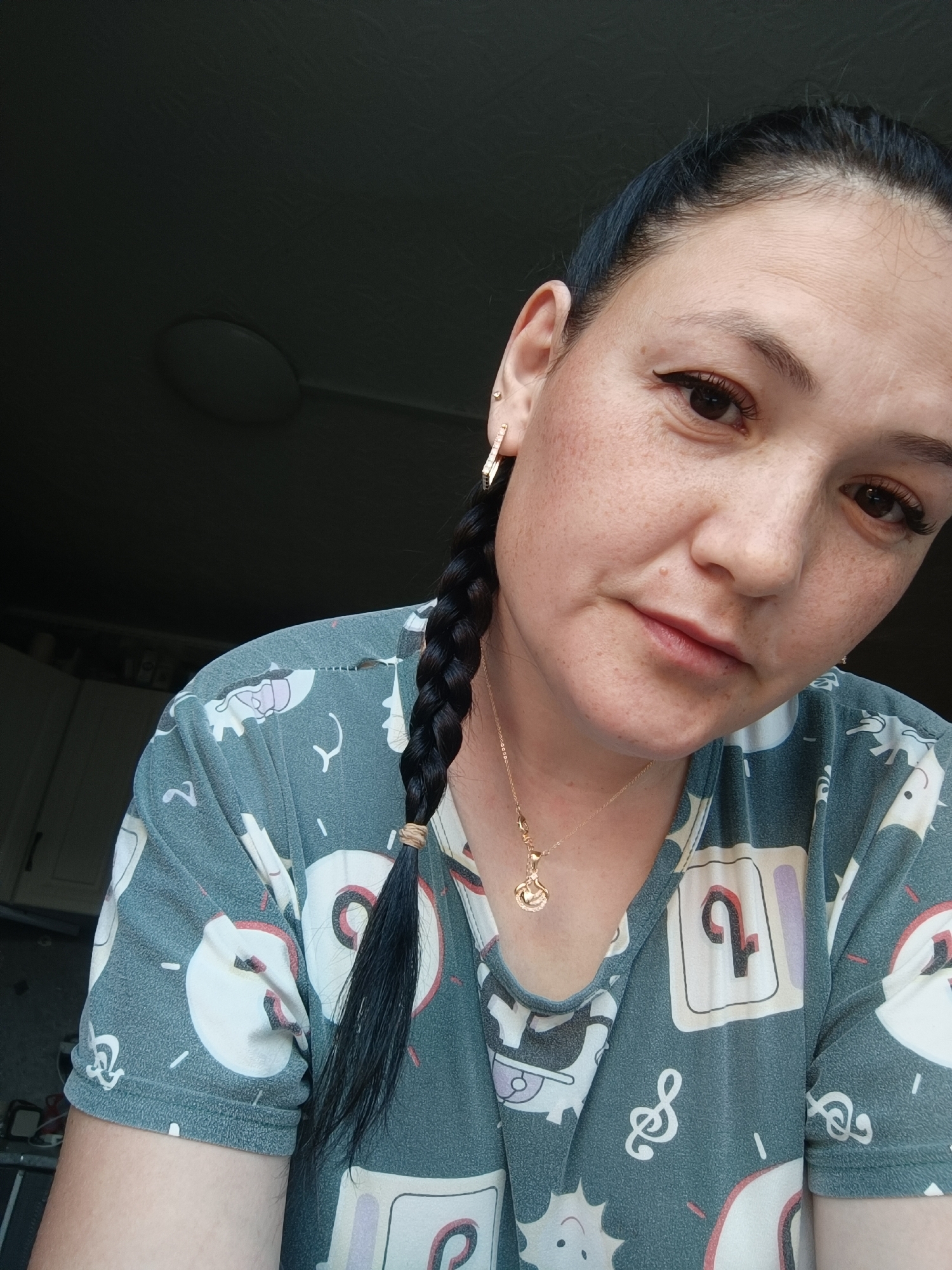A woman with dark hair styled in a braid and a gold necklace is seen from the chest up in a close-up, indoor shot. She is wearing a teal t-shirt with a repeating pattern of cartoon faces and musical notes. Her right ear is adorned with a geometric gold earring. The background is dark and out of focus, with a hint of a ceiling light fixture visible. There are kitchen cabinets and a countertop in the far left background. The scene appears to be in a domestic setting.