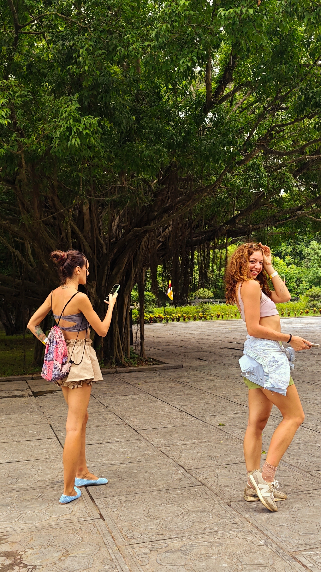 Two women are standing on a paved walkway in front of a large, ancient banyan tree with hanging roots. The woman on the left is facing away from the camera, holding a smartphone in her right hand and wearing a gray sports bra, a tan skirt, and blue polka-dot flats. She has a colorful backpack slung over her left shoulder. The woman on the right is facing the camera, smiling and touching her hair. She is wearing a pink crop top, a light blue denim jacket tied around her waist, light green shorts, and white sneakers with pink socks. The walkway is made of large, patterned stone tiles. In the background, there are lush green trees and foliage. A yellow and red flag is visible in the distance to the right of the banyan tree.