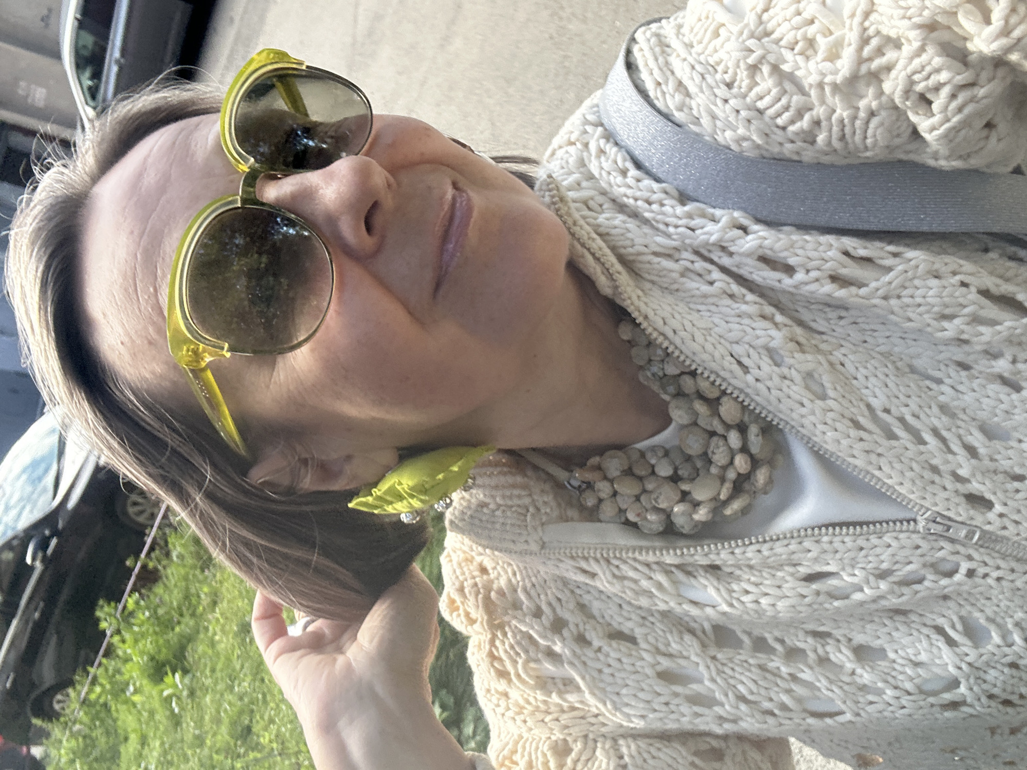 A woman with brown hair and sunglasses is shown from the chest up, facing forward and slightly to her right. She is wearing a cream-colored knitted sweater with an open zipper and a light-colored shirt underneath. A chunky necklace made of light-colored stones is visible, as are large, leaf-shaped earrings. A silver strap from a bag crosses her shoulder. In the background, out of focus, a dark-colored car is parked on a street next to a grassy area. The lighting suggests it is daytime.