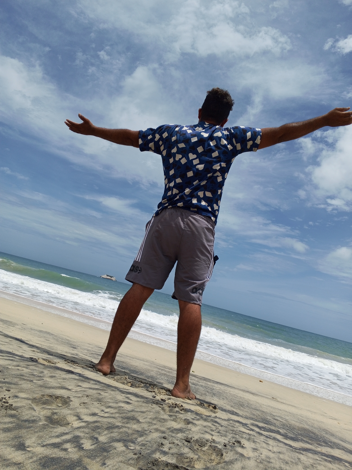 A man stands barefoot on a beach, arms outstretched, facing the sea. He wears a blue patterned shirt and gray shorts. The beach is sandy with dark streaks in the sand. Waves are visible crashing on the shore, and there's a boat in the distance on the sea. The sky is blue with scattered clouds. The location of Altagracia, Venezuela, is not verifiable.