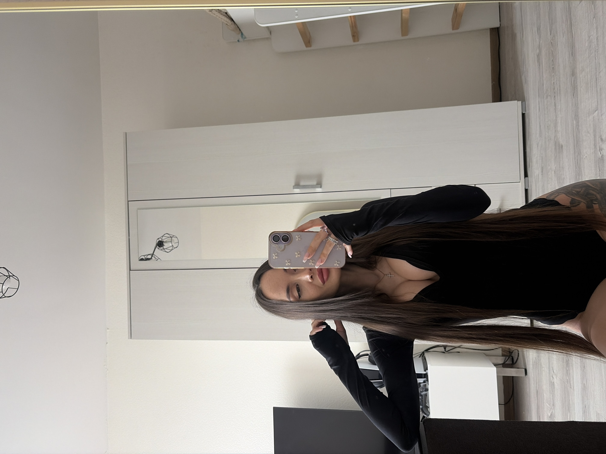 A woman with long brown hair, wearing a black long-sleeve top, is taking a mirror selfie with her phone. The background includes a white wardrobe with a mirrored door, a white wall with a decorative wire art of a dandelion clock, a white desk with a white monitor, and part of a grey upholstered chair. The scene is indoors, likely in an apartment or house. There is no indication of the location being in Saint Petersburg, Russia.