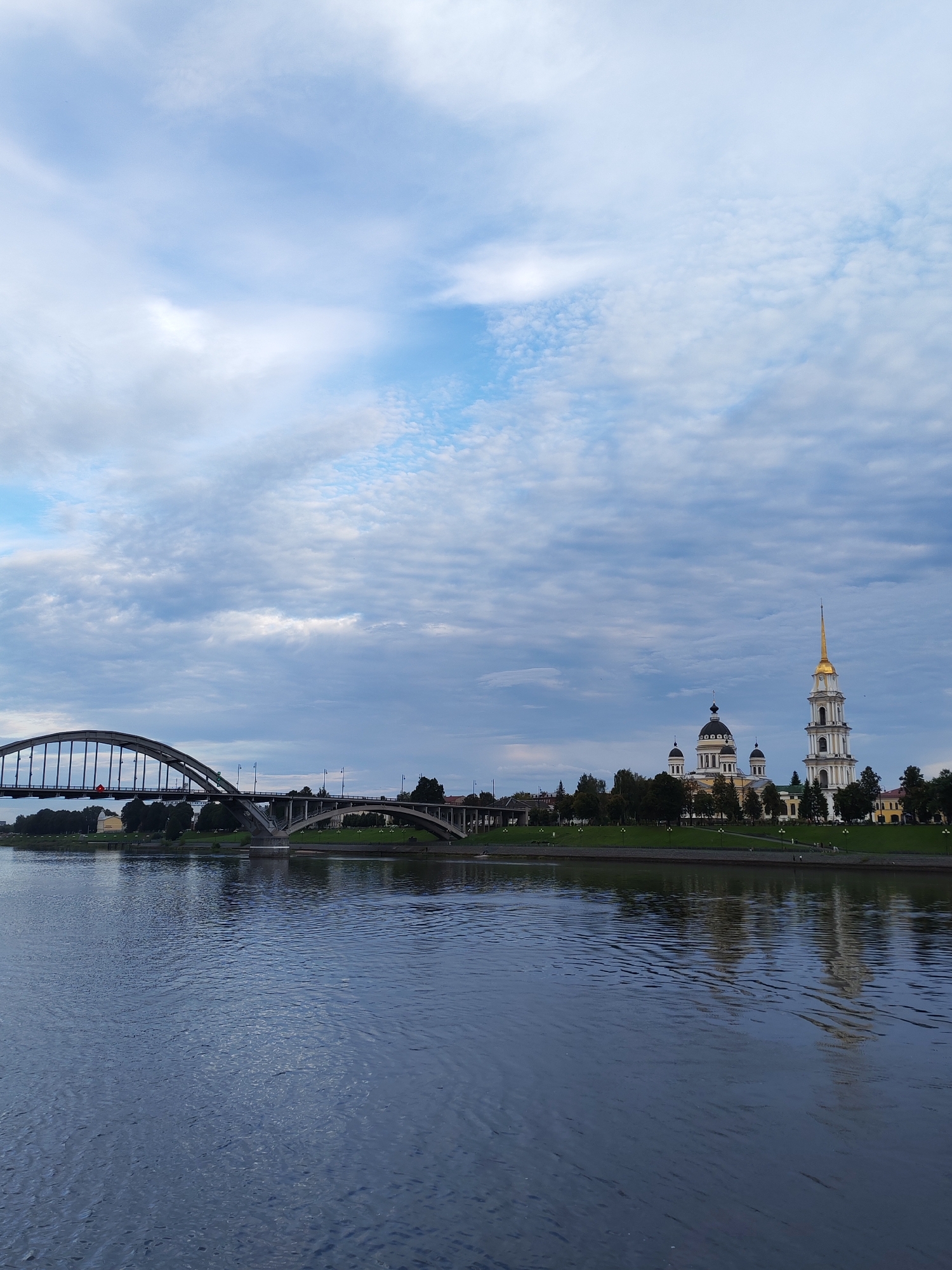 This image shows a wide, dark blue river in the foreground. On the far bank, a long, arched bridge spans the river, supported by several concrete piers. Beyond the bridge, a strip of green parkland stretches along the riverbank, dotted with trees and lampposts. In the distance, a complex of white and yellow buildings with ornate domes and a tall bell tower with a golden spire is visible. The sky above is a mix of blue and white clouds, with some darker grey clouds present. The reflection of the buildings and the sky is visible on the surface of the river. The scene appears to be a waterfront view of a city, possibly depicting a historical or religious landmark. The location is identified as Bol'shie Mkhi, Russia.