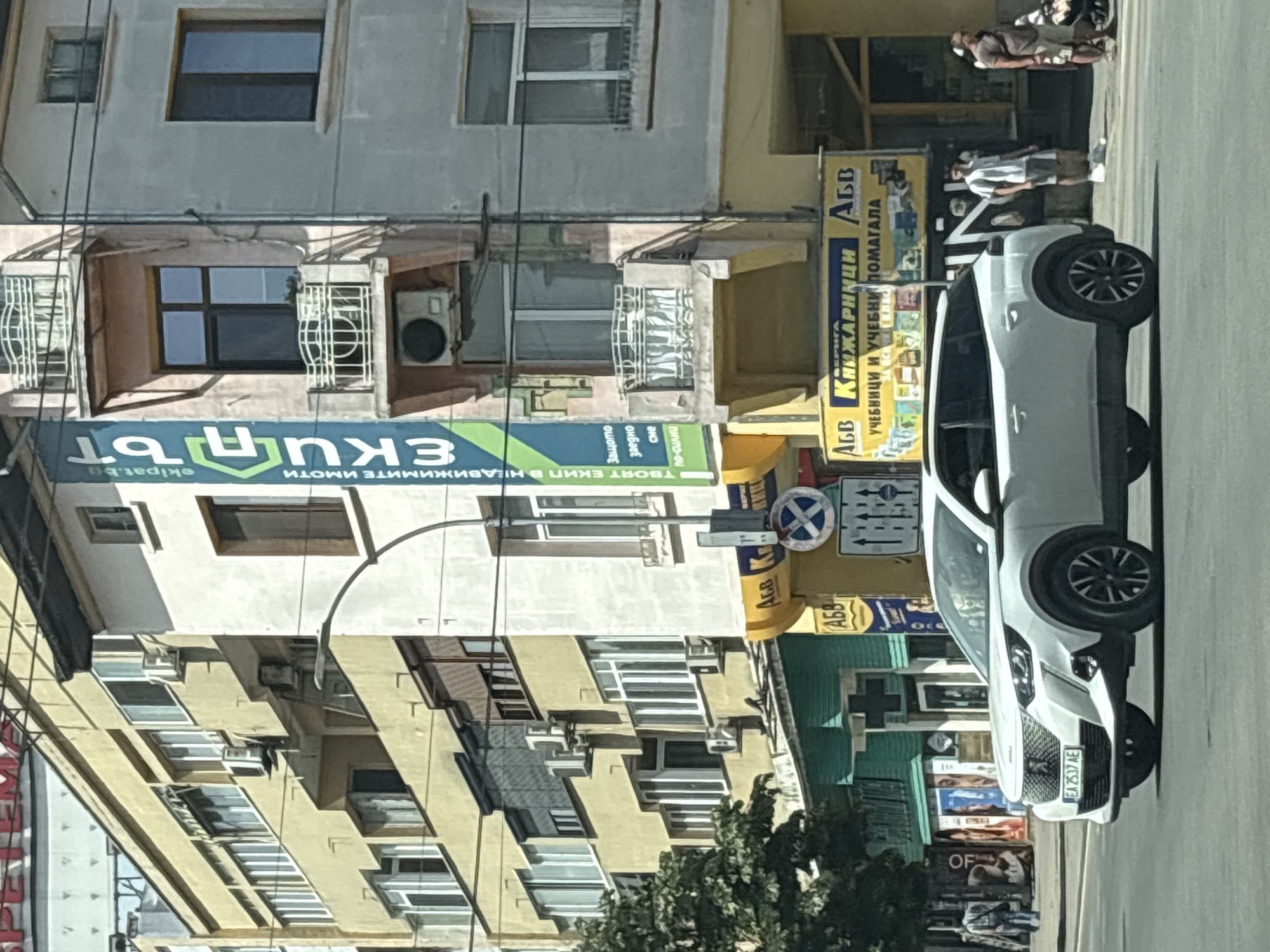 The image captures a street scene in Varna, Bulgaria, focusing on a white Peugeot 2008 SUV driving on a paved road. The vehicle's license plate reads "EA 2537 AE". In the background, there are multi-story buildings with a variety of architectural styles and colors, including yellow, pink, and light gray. One building features a prominent blue and green banner with text in Bulgarian. Several shops are visible at street level, including a bookstore with a yellow awning displaying "КНИЖАРНИЦИ" (Bookstores). Pedestrians are present on the sidewalk; one person in a white t-shirt and shorts stands near the bookstore, and another person in casual clothing walks with a stroller. The overall environment suggests an urban setting with commercial activity.