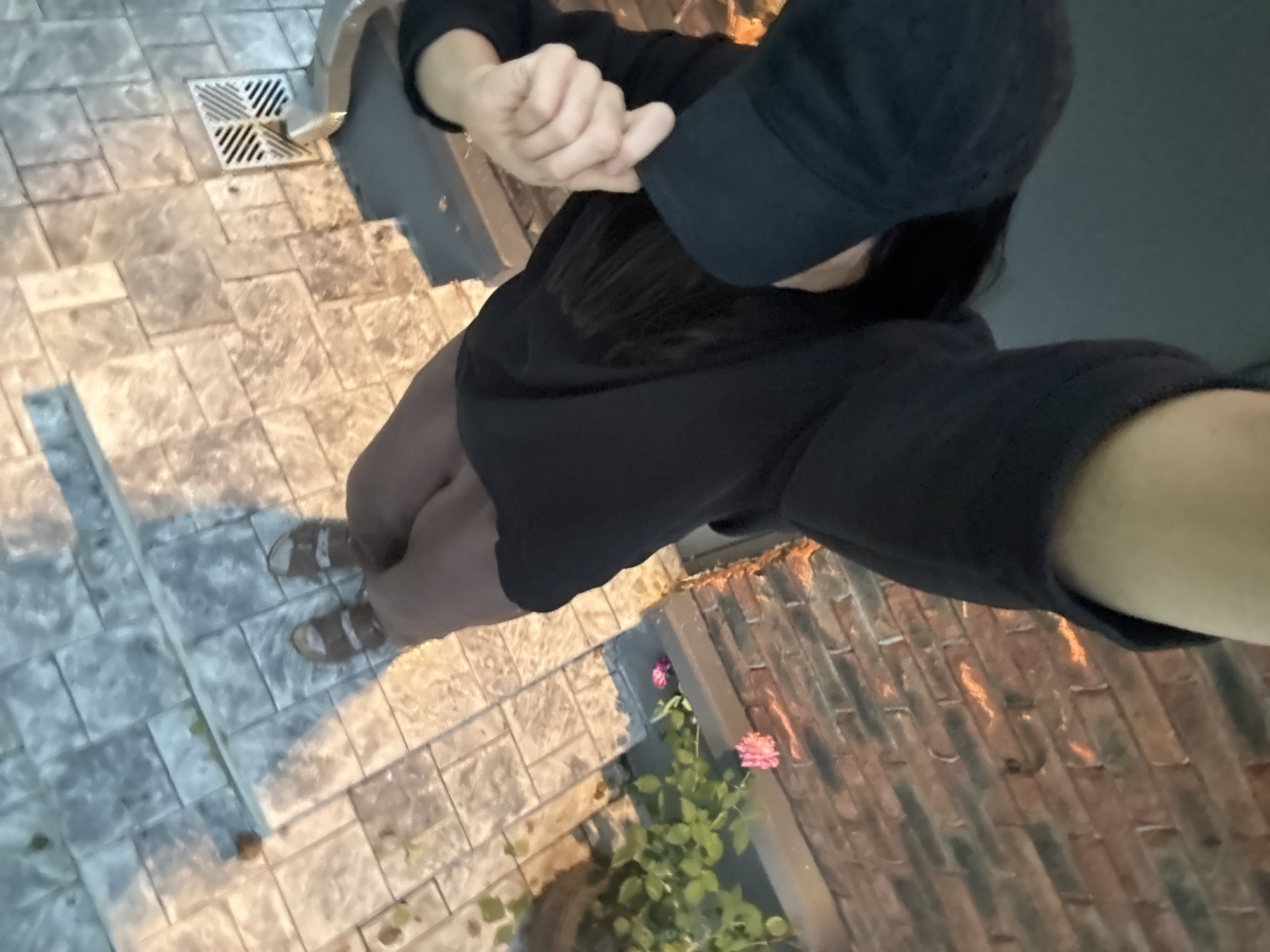 A person wearing a black baseball cap and a black sweatshirt stands on a paved area. The person's left hand is raised and clasped over their right wrist. The person is wearing brown leggings and brown sandals. In the background, there are pink flowers and green foliage. The paving stones are in a brick pattern and appear to be wet. To the left of the person, there is a metal grate. The scene is dimly lit, possibly at dusk or night. This image was taken in the city of Chaikovsky, Russia.