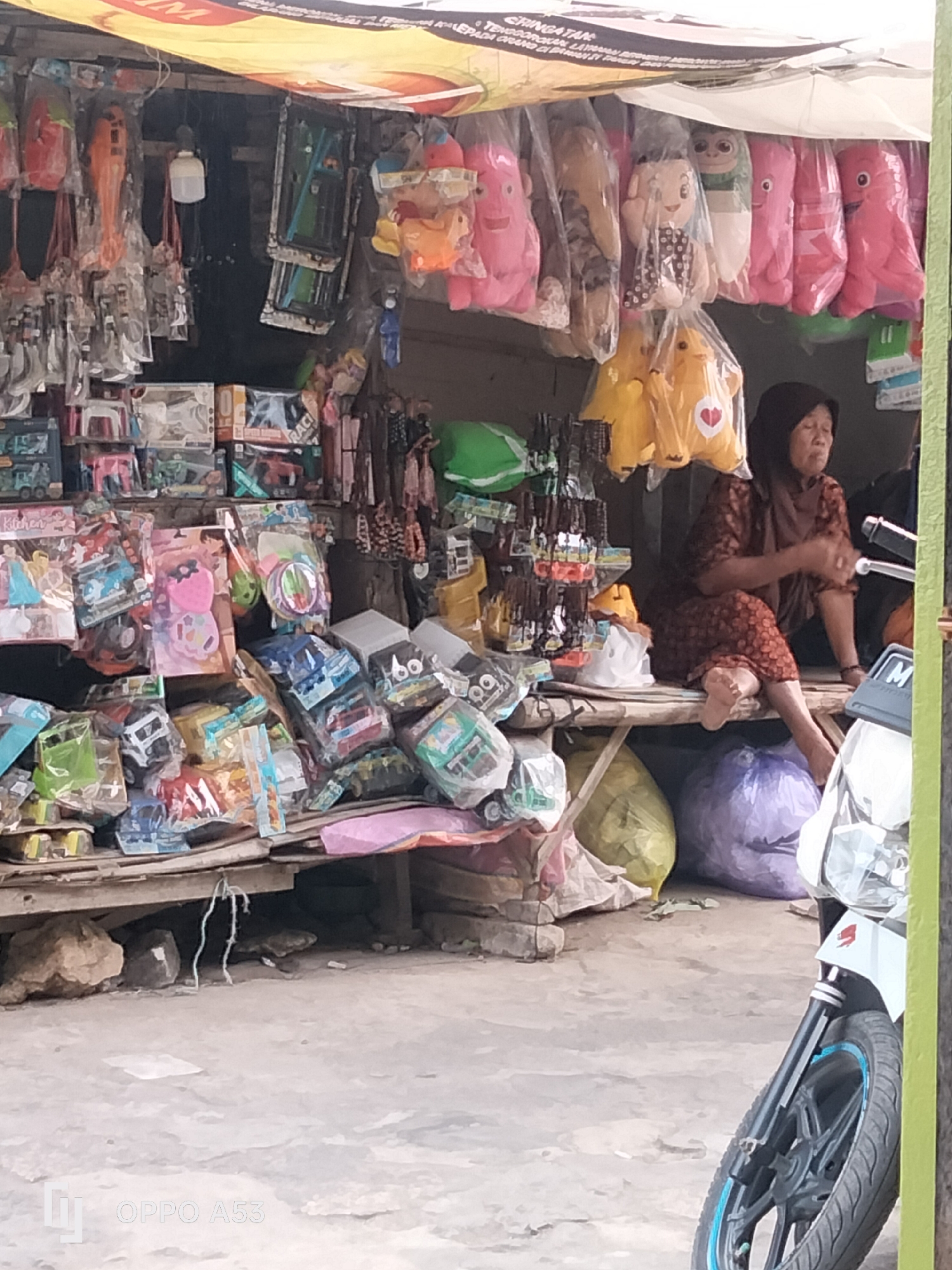 A woman in a brown and maroon patterned headscarf and dress sits on a wooden platform in front of a stall selling toys and various merchandise. The stall has a yellow awning and is cluttered with a variety of items, including stuffed animals, toy cars, and other packaged goods. The woman appears to be resting, with her eyes closed. Behind her, several large sacks of goods are visible, one yellow and one purple. In the foreground, the front of a white motorcycle with black spokes and blue trim is visible, along with a portion of its handlebars. The ground is paved with concrete. The overall scene suggests a small, informal marketplace.