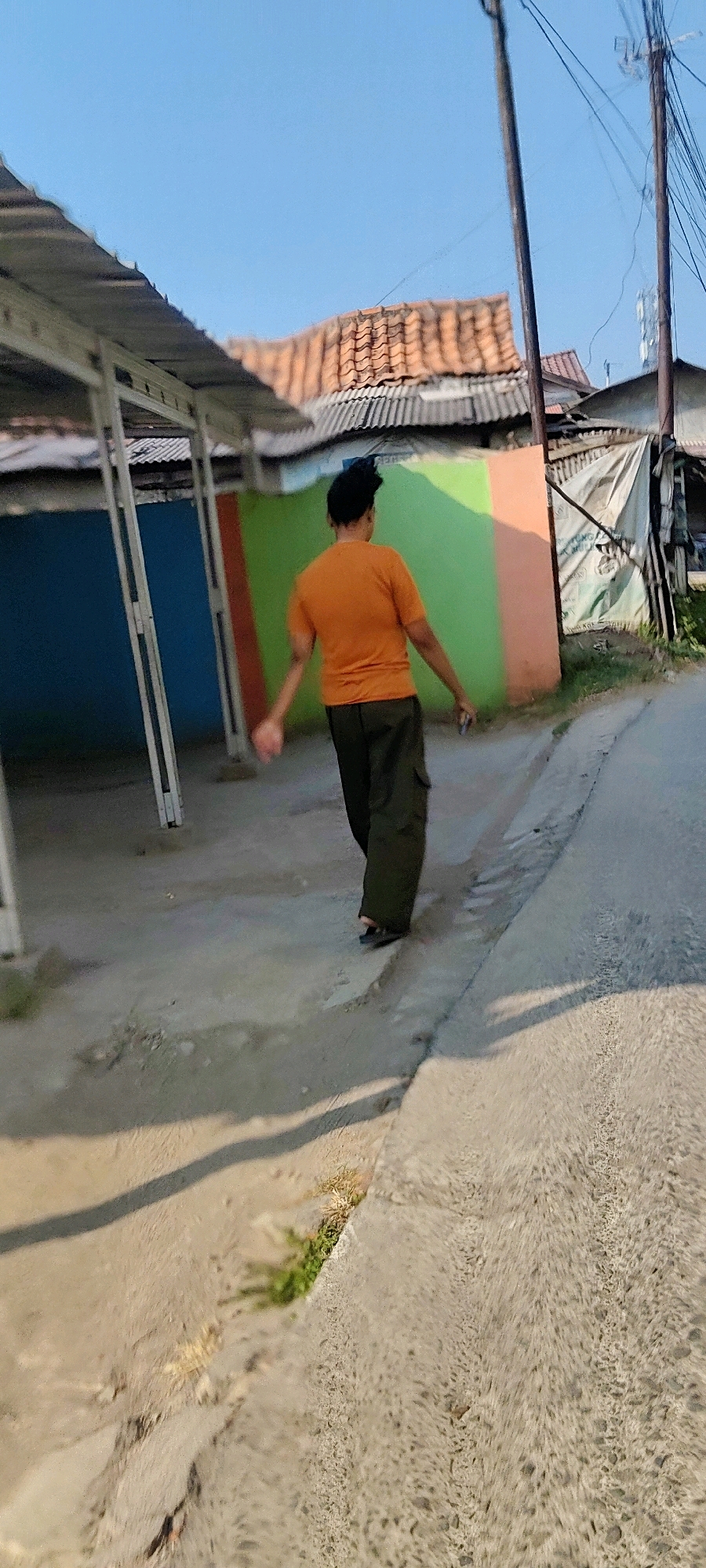 In Mekarmukti, Indonesia, a person wearing an orange t-shirt and dark green cargo pants walks away from the camera on a paved area next to a dirt path. Behind the person are buildings with tiled roofs and walls painted in vibrant colors, including blue, green, and orange. Utility poles with numerous wires are visible against a clear blue sky. The scene appears to be an outdoor, possibly residential or commercial, area in a village or town.