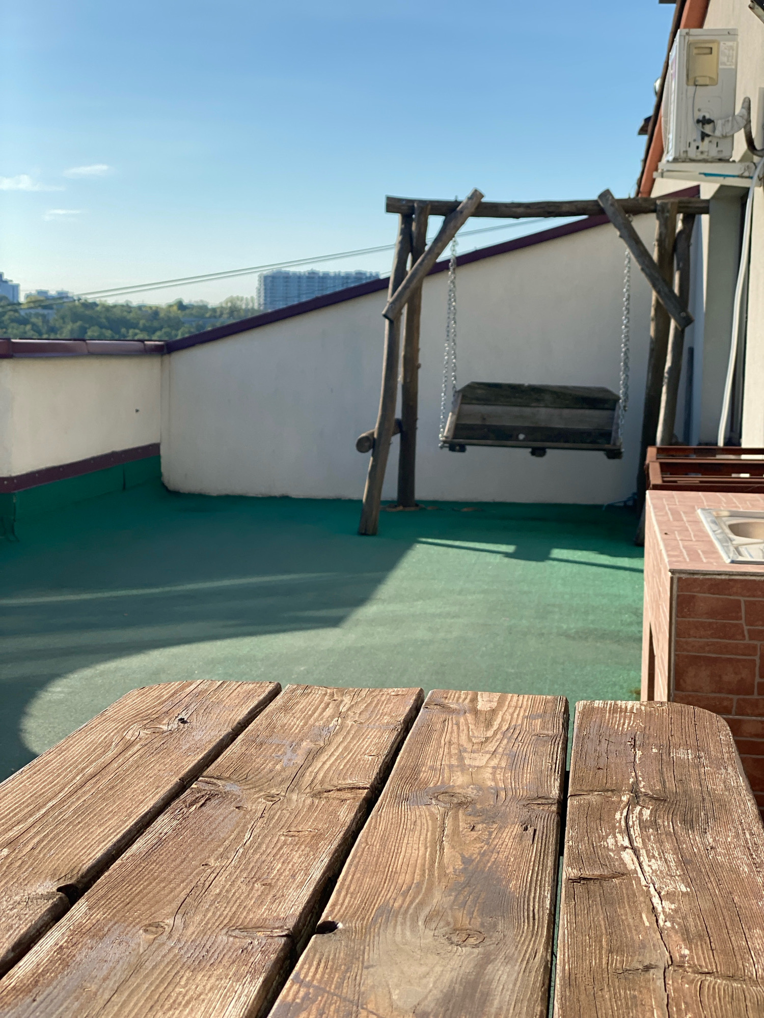 The image depicts a rooftop patio with a wooden swing. In the foreground, a weathered wooden table is visible. The patio floor is covered in green turf. In the background, a white wall with a purple trim runs along the edge of the patio. Beyond the wall, a cityscape with buildings and trees can be seen under a clear blue sky. An air conditioning unit is mounted on the wall of a building to the right. A brick structure with a sink is also partially visible on the right side of the image. The scene appears to be in Chișinău, Moldova, though no specific landmarks are identifiable. There are no people or animals present.