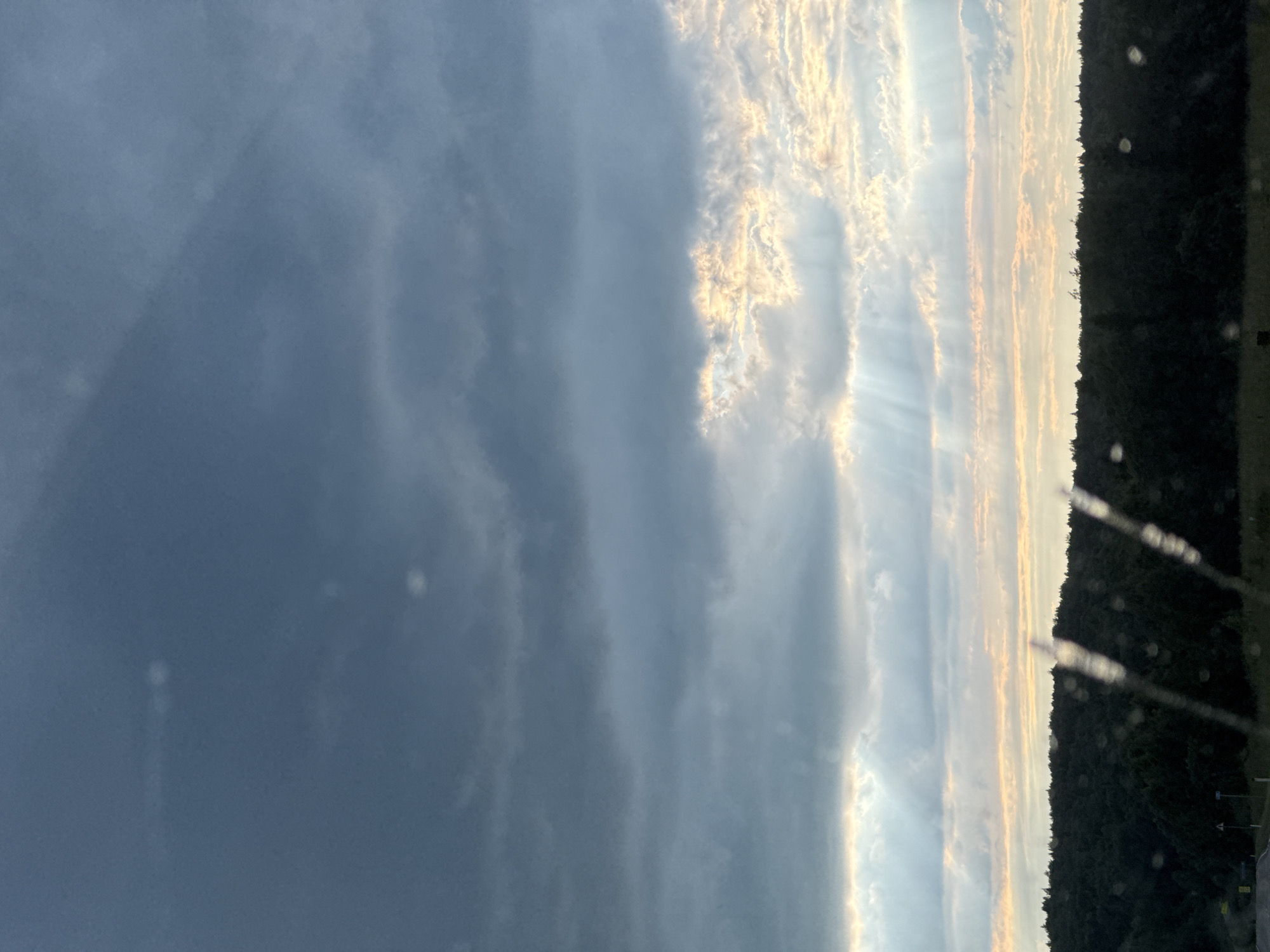 The image captures a dramatic sky over a densely forested area. Dark, heavy clouds dominate the upper portion of the frame, suggesting an approaching storm or the aftermath of one. Shafts of light, known as crepuscular rays, pierce through gaps in the clouds lower in the sky, illuminating patches of lighter, broken clouds with warm, golden hues. Below the sky, a dark, silhouetted line of trees forms a dense forest that stretches across the horizon. The foreground is blurred, with vertical streaks that appear to be water droplets or streaks on a window, hinting that the photograph may have been taken from inside a vehicle. The environment appears to be natural and rural. There are no people or specific man-made structures clearly visible in the main scene, although a faint road and signs are present in the far lower left corner. The overall mood is one of natural power and atmospheric beauty. The location is specified as Большая Уса, Russia.
