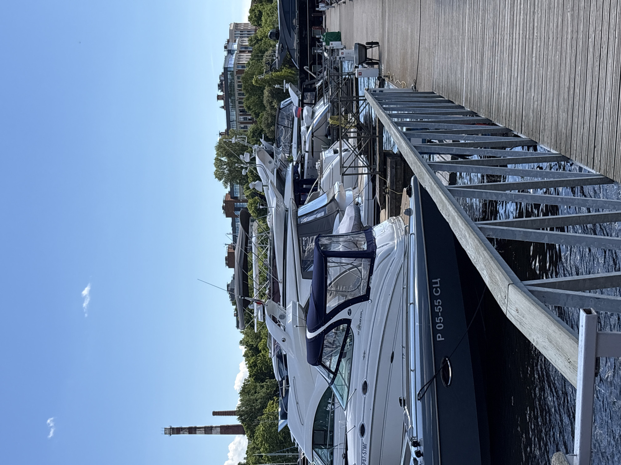 The image captures a sunny day at a marina in Saint Petersburg, Russia, likely along the Neva River or one of its tributaries, given the industrial chimneys in the background, which are characteristic of the city's historical industrial zones. Several yachts and smaller boats are docked along a wooden pier. In the foreground, a white and grey motorboat is prominently featured, with a blue canvas cover over its cockpit. The boat displays the registration number "P 05-55 CU" on its hull. Beyond this boat, other vessels of varying sizes are moored, including larger white yachts with covered seating areas. The background reveals a skyline dominated by green trees, followed by multi-story buildings with modern architecture and a distant industrial chimney stack. The sky is a clear, bright blue with a few wispy clouds. The scene depicts a typical recreational boating environment within an urban setting.