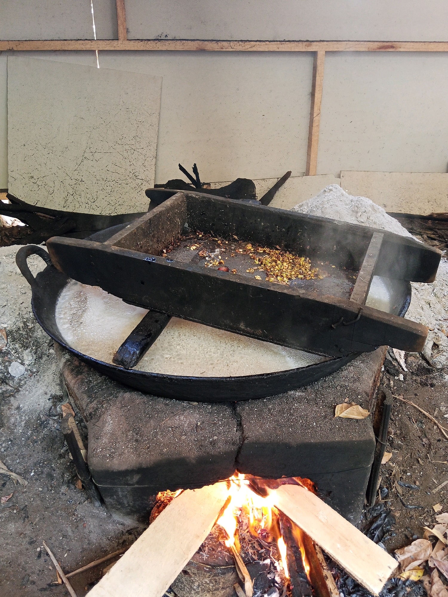 In Langgongsari, Indonesia, a cooking scene unfolds outdoors under a rudimentary shelter. A large, black cast-iron wok, set on a brick stove with an open fire below, is filled with a bubbling, light-colored liquid. Above the wok, a wooden apparatus, possibly for sifting or drying, holds a pile of small, yellowish-brown objects, some of which appear to be seeds or beans. Steam rises from the wok, indicating the cooking process is active. The surrounding ground is composed of dirt and ash, with scattered dry leaves. The shelter's walls are made of large, light-colored panels, possibly plaster or cardboard, supported by wooden beams. The overall impression is one of a traditional, possibly rural, food preparation method.