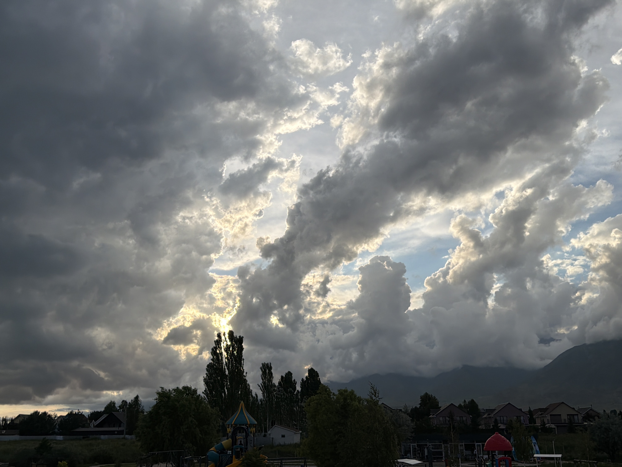 The image depicts a dramatic sky filled with dark, ominous clouds with breaks of bright sunlight peeking through, creating a dynamic contrast. In the foreground, a suburban landscape is visible, featuring houses with red-tiled roofs and pitched gables, nestled among trees. A playground structure with colorful elements, including a red dome-shaped slide, is present. The environment suggests a residential area on the outskirts of the city of Cholpon-Ata, Kyrgyzstan, with mountains visible in the distance. The overall scene captures a moment of changing weather, possibly before or after a storm.