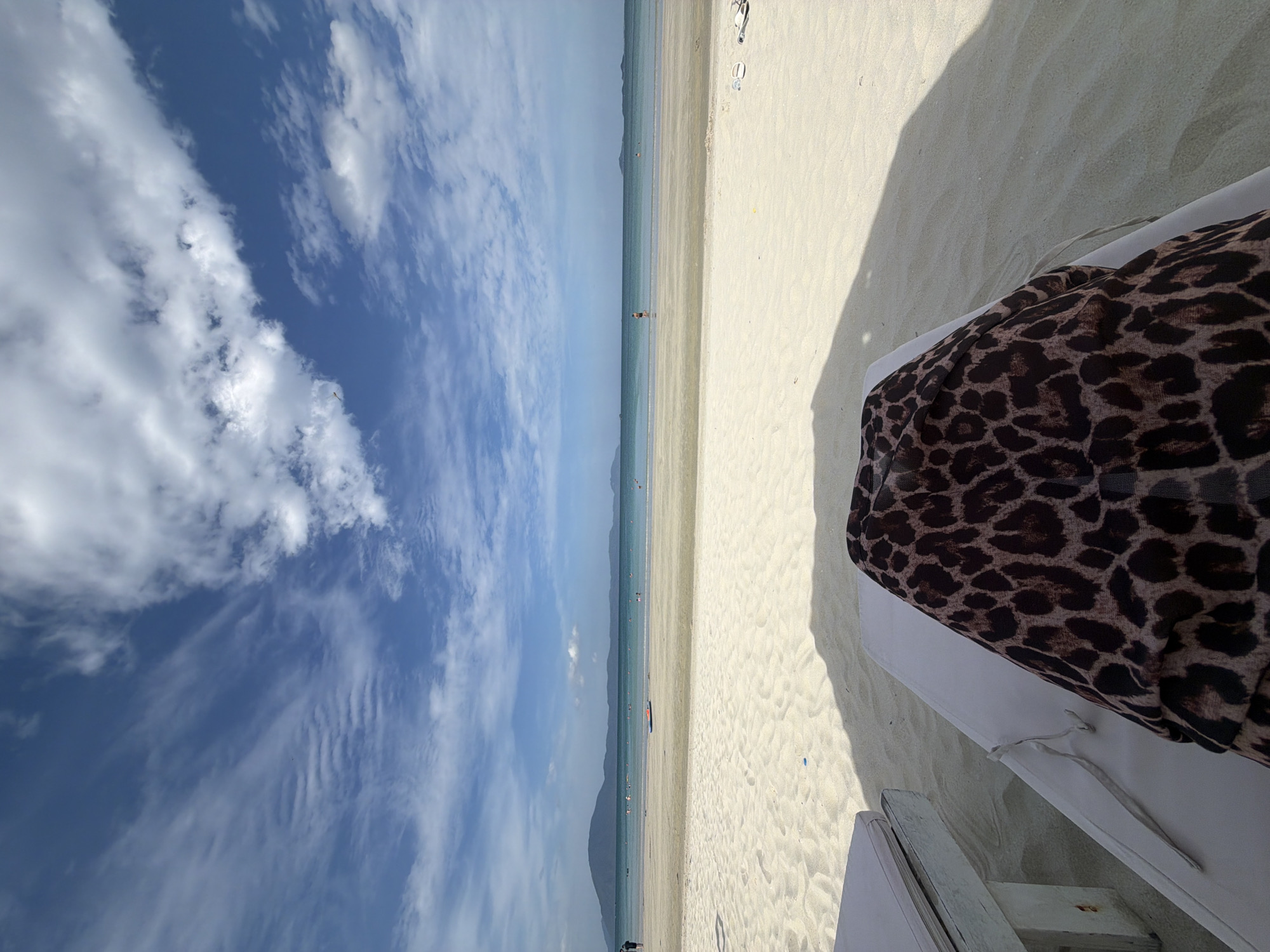 The image depicts a scene at a beach in Đông Ninh Hòa, Vietnam. The foreground shows a close-up of a beach chair with a leopard-print fabric draped over it. To the left, another beach chair is partially visible, with a white cushion and a weathered white wooden frame. The sand is light-colored and appears smooth, with shadows cast from the chairs and possibly an umbrella.
In the midground, a wide expanse of white sand stretches towards the ocean. Several individuals are scattered across the sand and in the shallow water. Some are wading or swimming, while others are standing or walking. A bright blue and orange inflatable object is visible in the water.
The background features a calm, turquoise ocean that meets a hazy horizon. The water is relatively shallow near the shore, with gentle waves. In the far distance, a line of mountainous islands can be seen against the horizon. The sky is a vibrant blue, filled with scattered white clouds, some of which are wispy and elongated, characteristic of cirrus clouds, while others are more cumulus in form. The overall impression is a serene beach environment on a clear day.