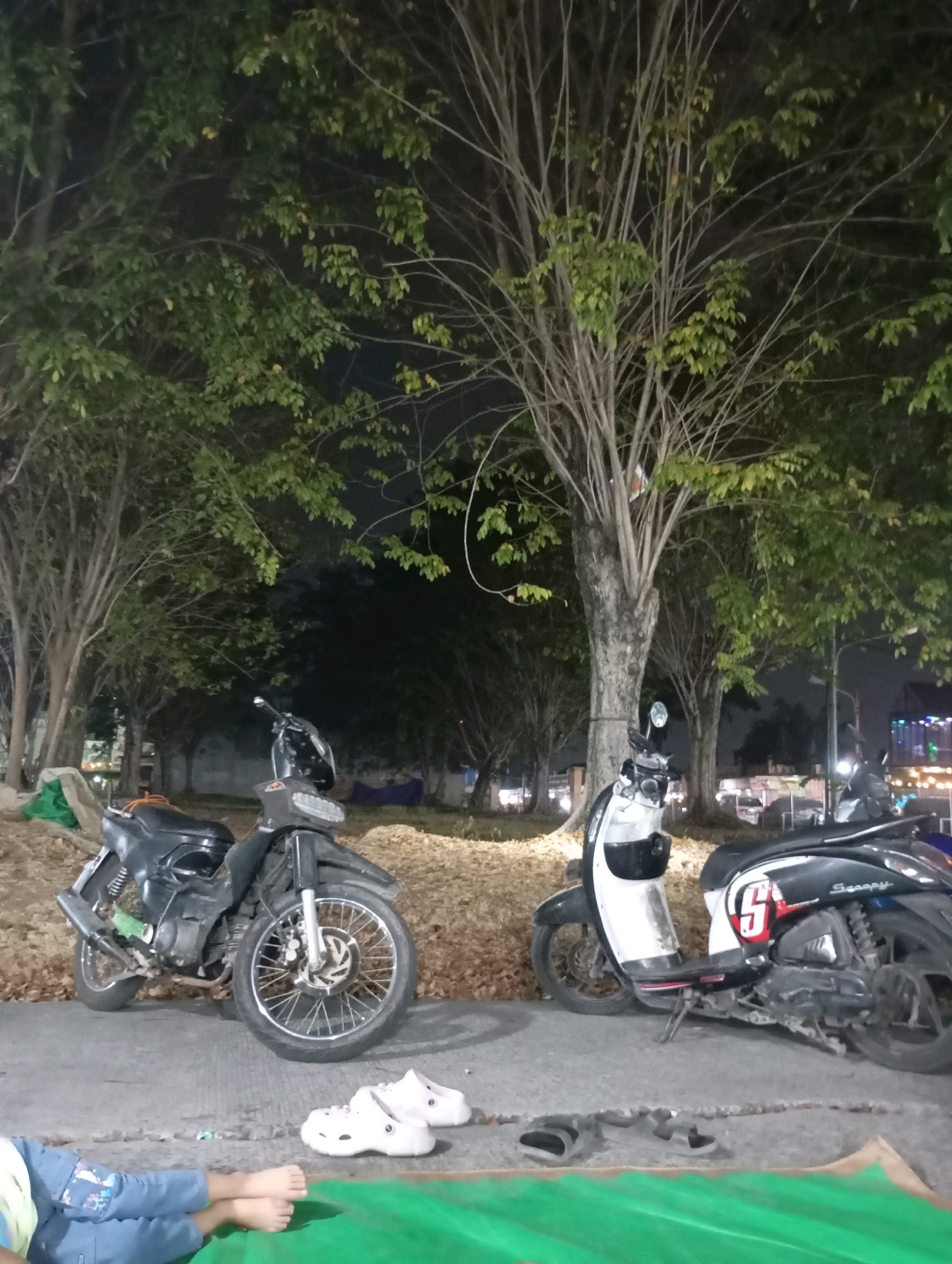 In Babelan, Indonesia, at night, two scooters are parked on a paved surface next to a grassy area with fallen leaves. A black scooter with a silver front wheel is on the left, and a white and black scooter with a red "5" sticker and "Scoopy" written on it is on the right. A person's legs, wearing blue pants with a cartoon pattern, are visible in the lower left corner, lying on a green tarp. Two pairs of white clogs are placed near the scooters. The background features trees with sparse foliage and a dimly lit street with buildings and illuminated signs.